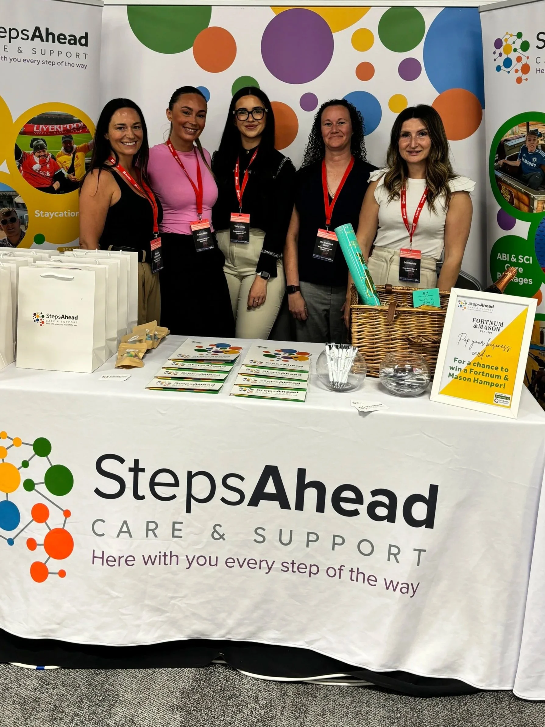 Group of five women standing behind a booth with promotional materials for Steps Ahead Care & Support at a convention or event. The booth has a white tablecloth with the company logo and slogan, and various pamphlets, signs, and a basket with rolled-up items on display. Background features colorful circular graphics and banners.