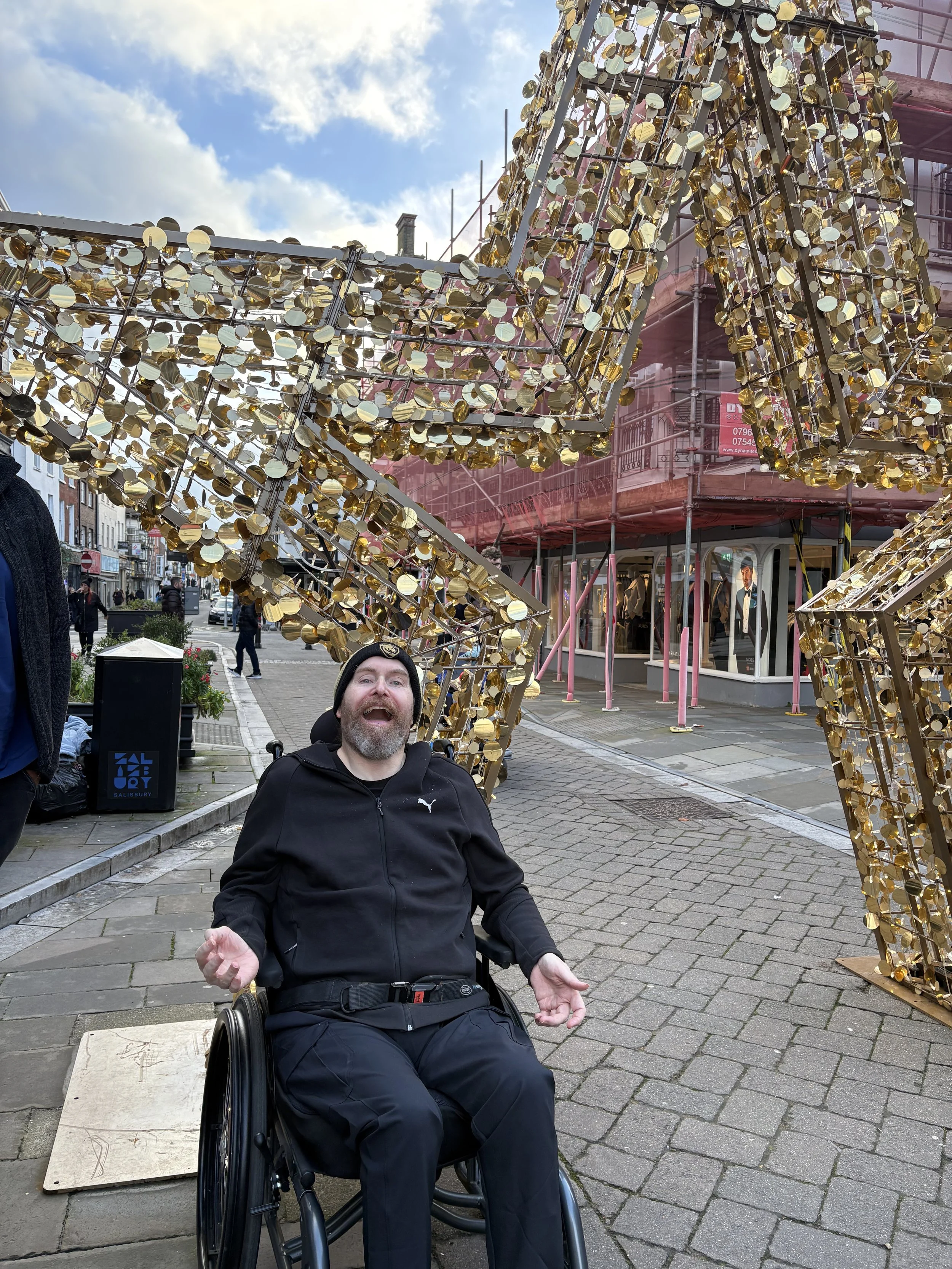 A man with a beard and black jacket sitting in a wheelchair, smiling and gesturing with his hands, in front of a large, illuminated, gold-colored decorative structure on a city sidewalk.