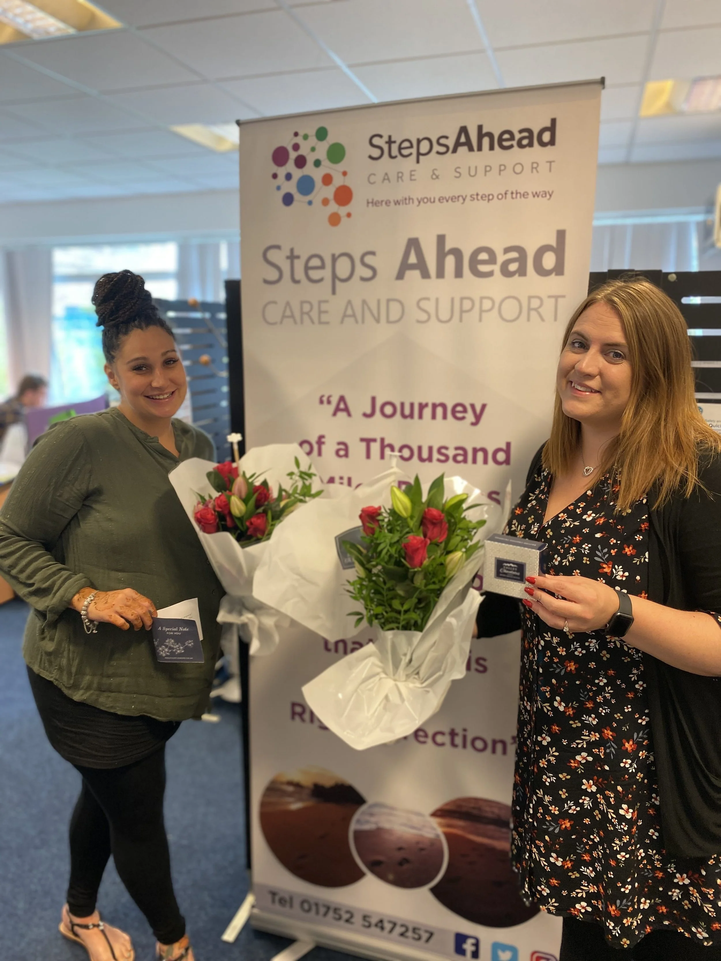 Two women standing next to a banner and holding bouquets of flowers at a charity event.