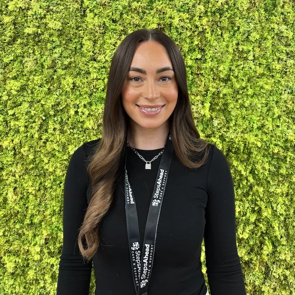 Young woman with long brown hair, wearing a black top and a lanyard, smiling in front of a green foliage wall.