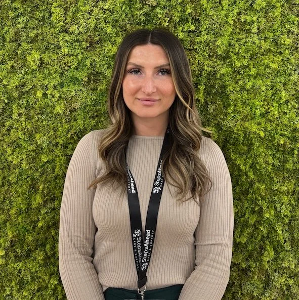A woman with long wavy hair wearing a beige ribbed sweater and a black lanyard with white text, standing against a green leafy background.