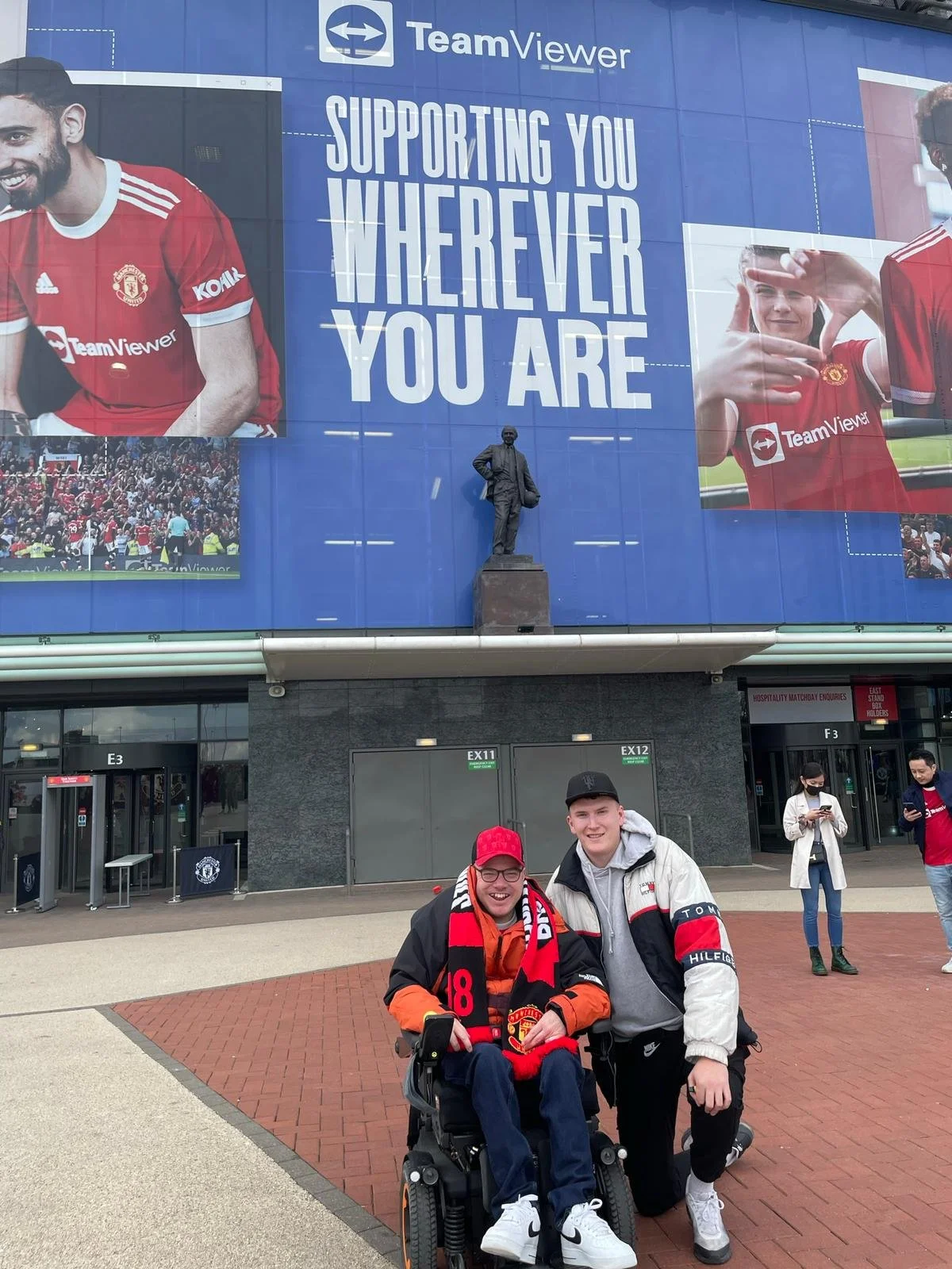 Two young men smiling and posing for a photo outside a sports stadium. One is seated in a wheelchair wearing a red Manchester United hat and scarf, the other stands beside him wearing a black cap and a white jacket. Behind them is a large stadium entrance with people walking and a billboard with Manchester United fans and a Chelsea statue.