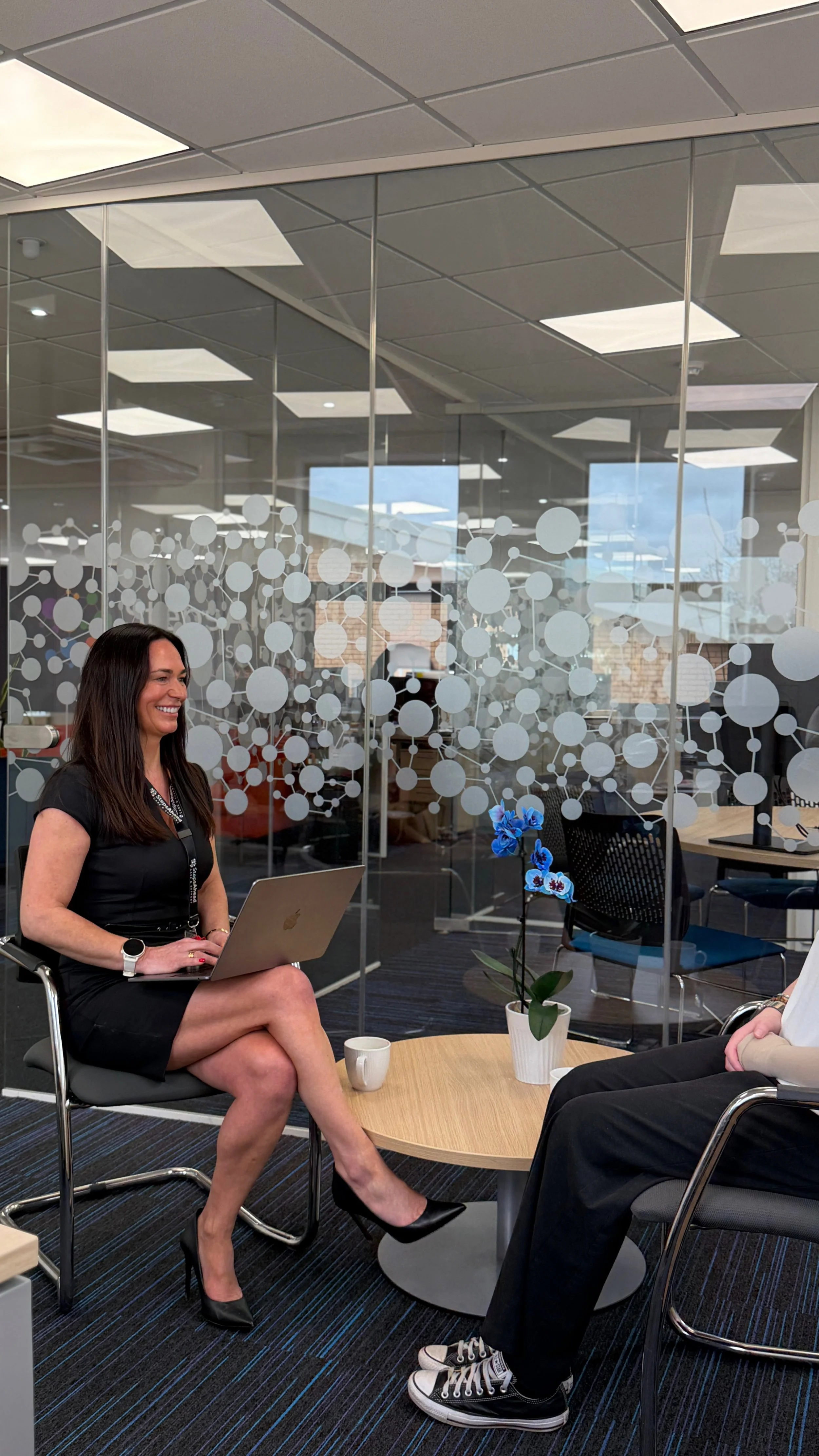 A woman with long dark hair, wearing a black dress and black high heels, sitting in an office, smiling, holding a laptop on her lap. She sits across from another person, partially visible, wearing black pants and sneakers. Between them is a small wooden table with a blue orchid in a white pot and two white coffee mugs. The background shows a modern office with glass walls and ceiling lights.