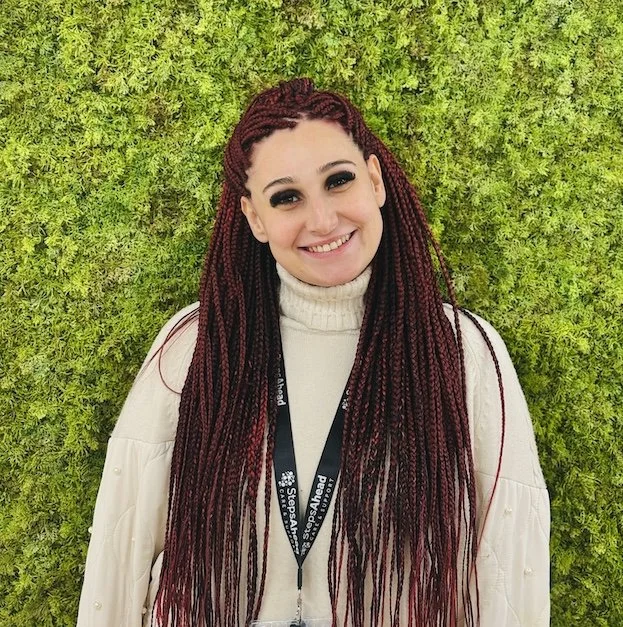 A woman with long red braided hair smiling, standing in front of a green leafy wall, wearing a white turtleneck and a black lanyard.