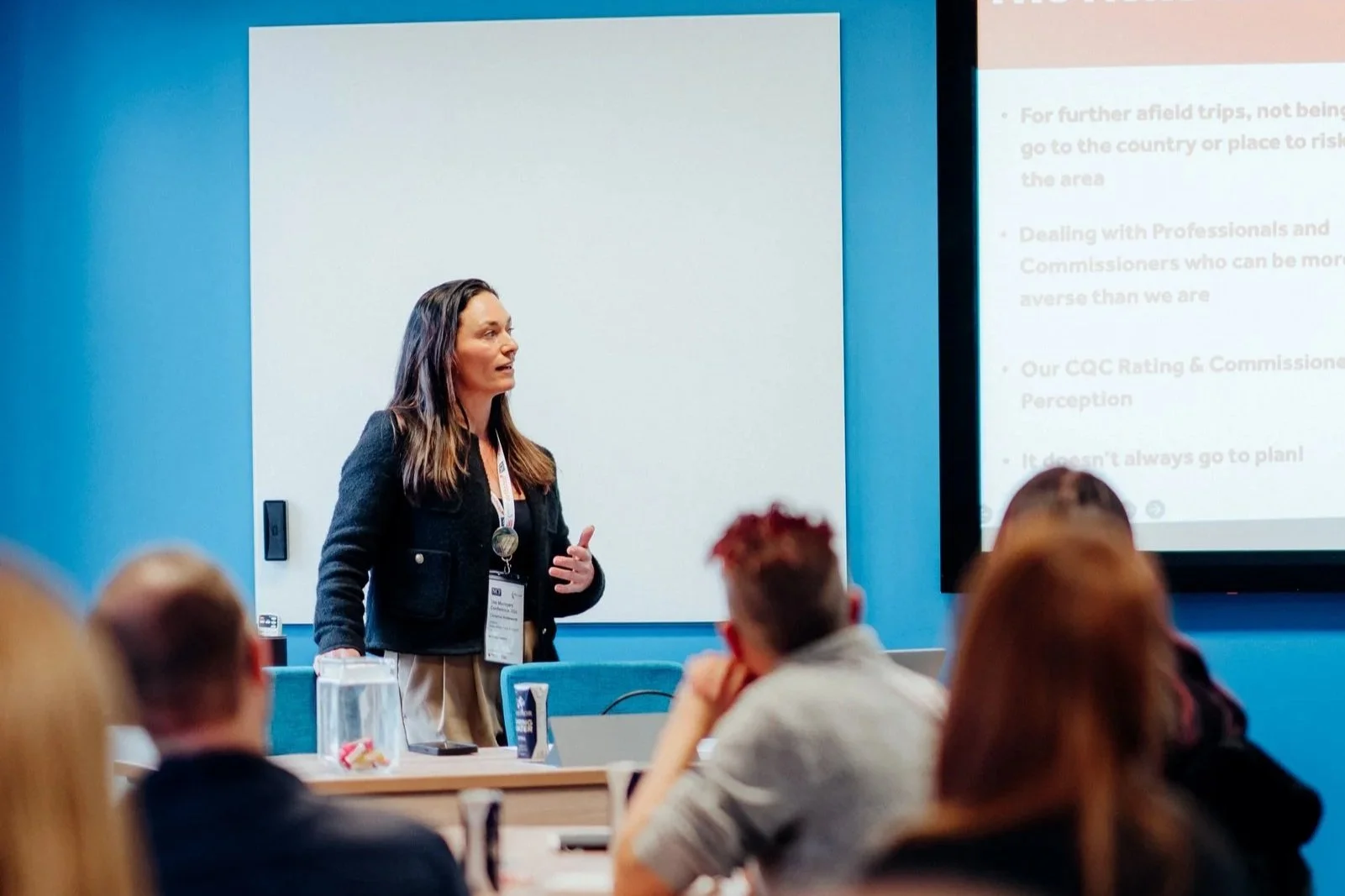 Woman giving a presentation in a conference room with several attendees watching her. She is standing near a presentation screen with text, and the room has a blue wall.
