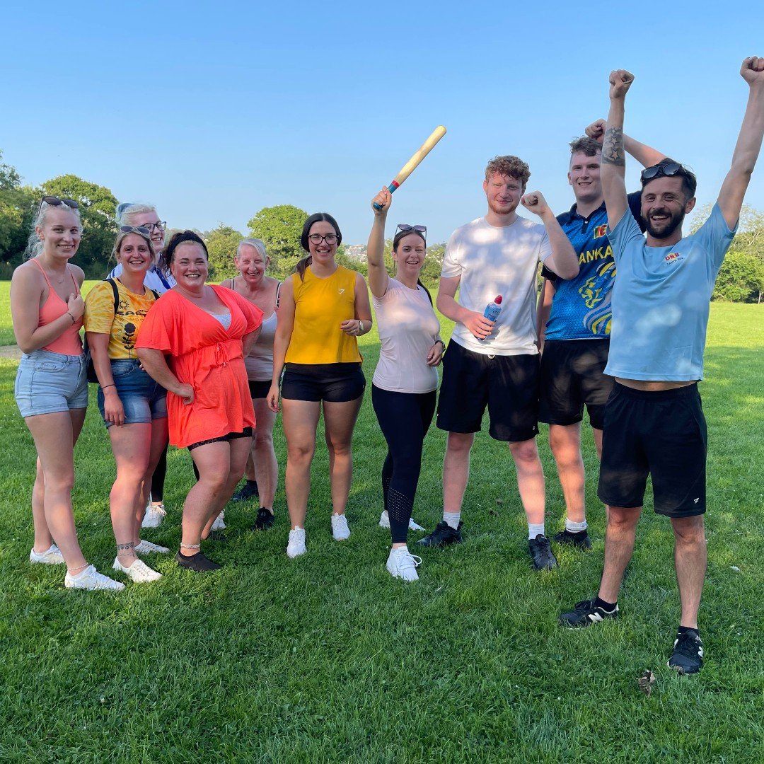 A group of ten people standing on a grassy field under a clear blue sky, celebrating with smiles and raised arms, some holding water bottles, during a sunny day.