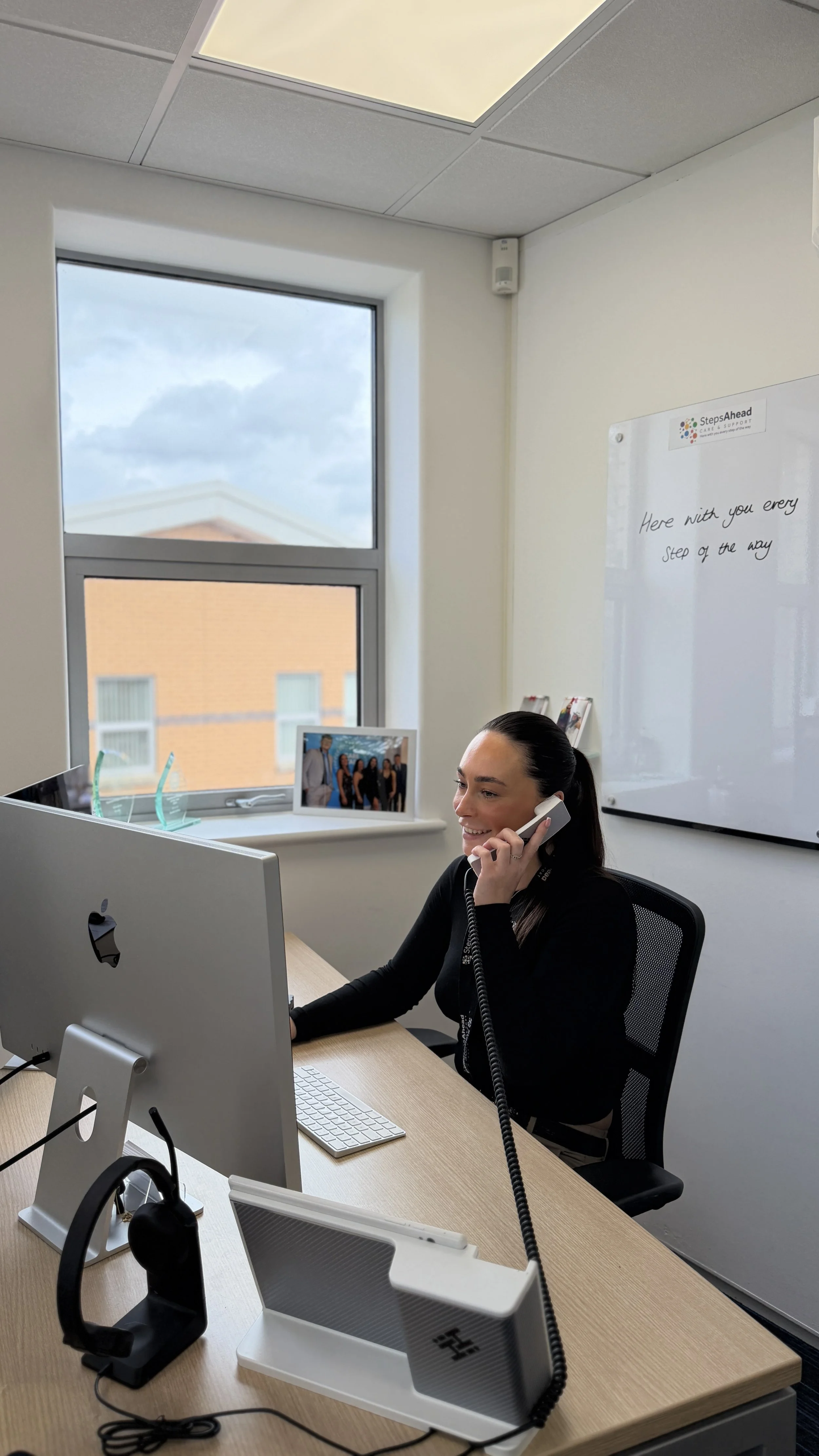 A woman sitting at a desk in an office, talking on a landline phone, with a computer monitor, keyboard, and headset on the desk. A window behind her shows a cloudy sky and a building, and a whiteboard on the wall has the message "Here with you every step of the way."