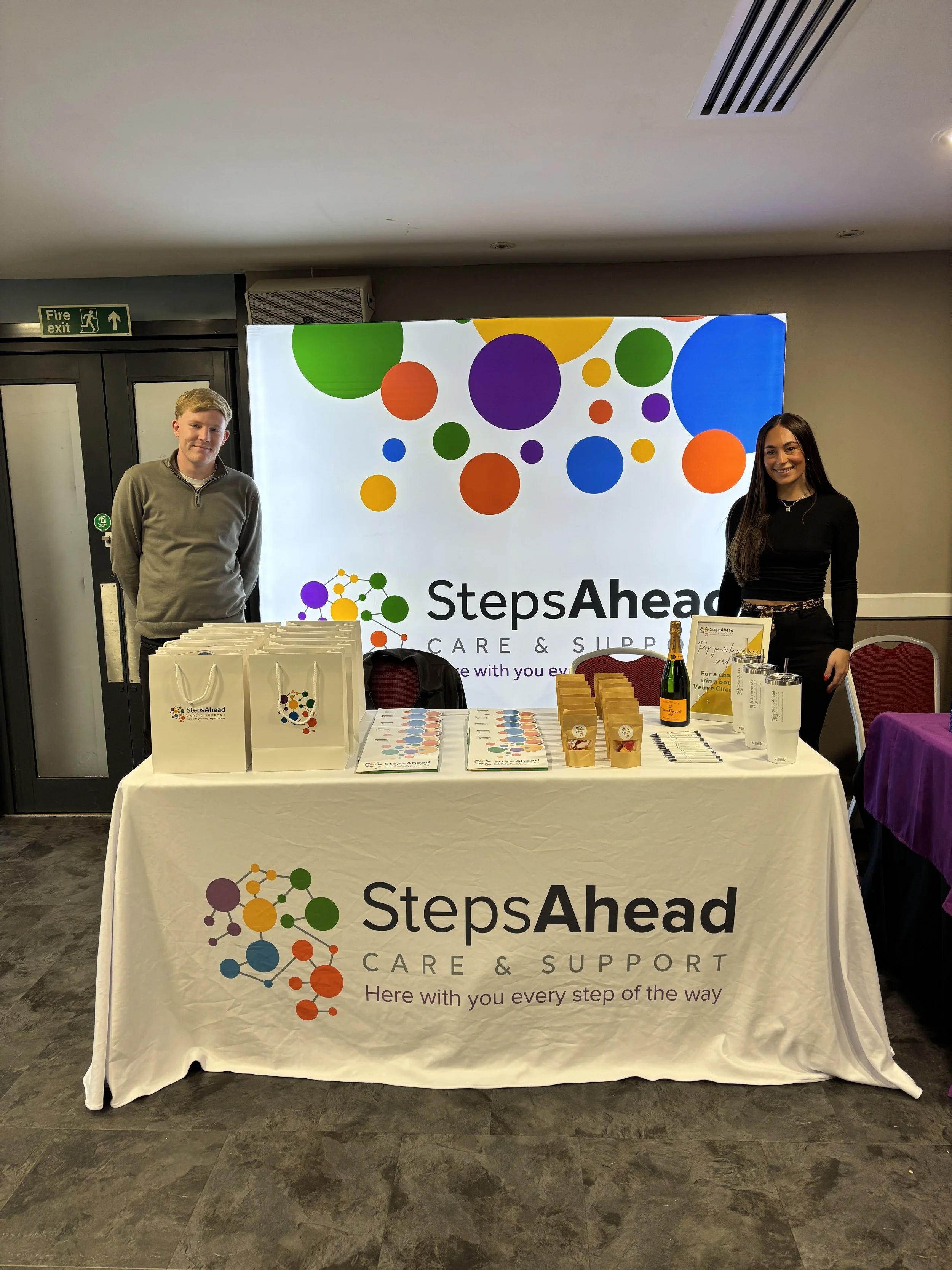 A promotional table for StepsAhead Care & Support with a large logo and slogan on a white tablecloth. Behind are a man in a grey sweater and a woman in black, standing next to a colorful backdrop with the same logo. The table has brochures, small bags, a framed sign, and a bottle, with a purple-covered table on the right.