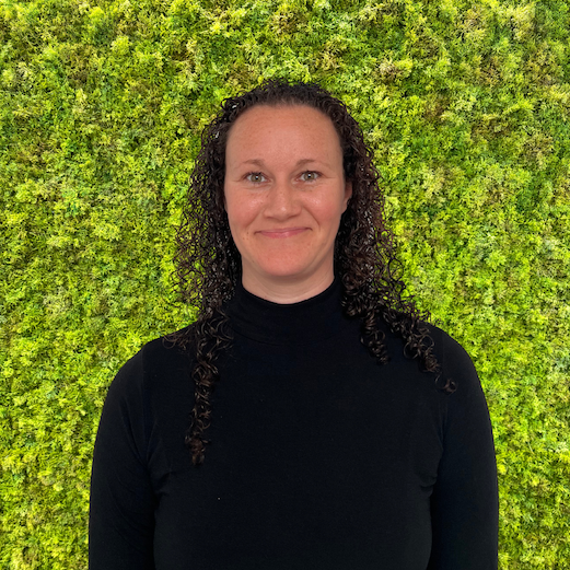 A woman with curly dark hair wearing a black turtleneck sweater standing in front of a green leafy wall.