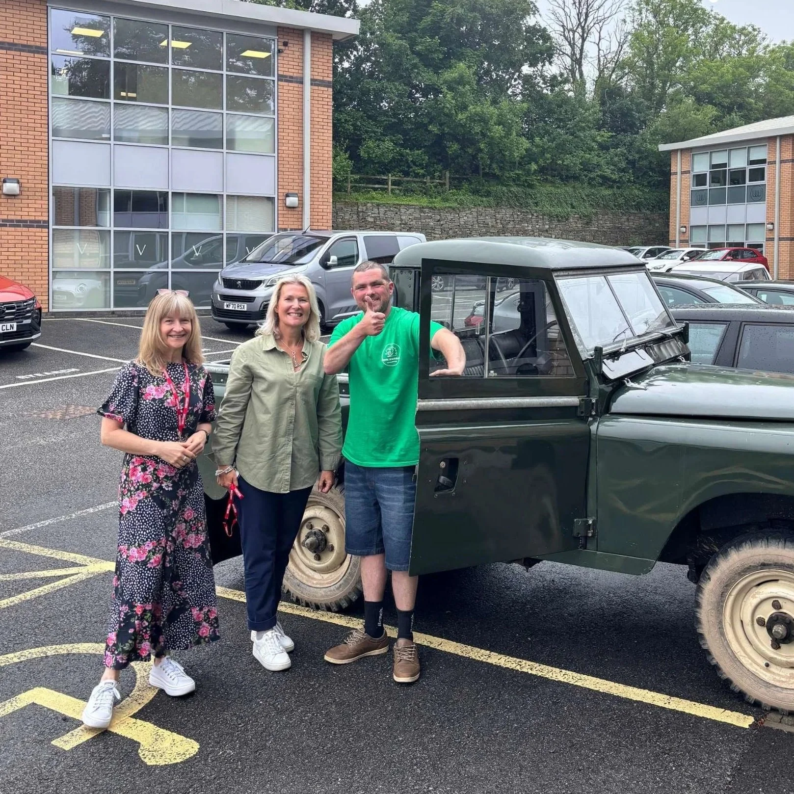Three people standing next to a vintage military jeep in a parking lot. The person on the right is giving a thumbs up. The other two are smiling, with a woman on the left wearing a polka-dot dress with floral pattern and white sneakers, and a woman in the middle wearing a green jacket and dark pants. There are several modern cars and a brick building with large windows in the background, and green trees behind the building.