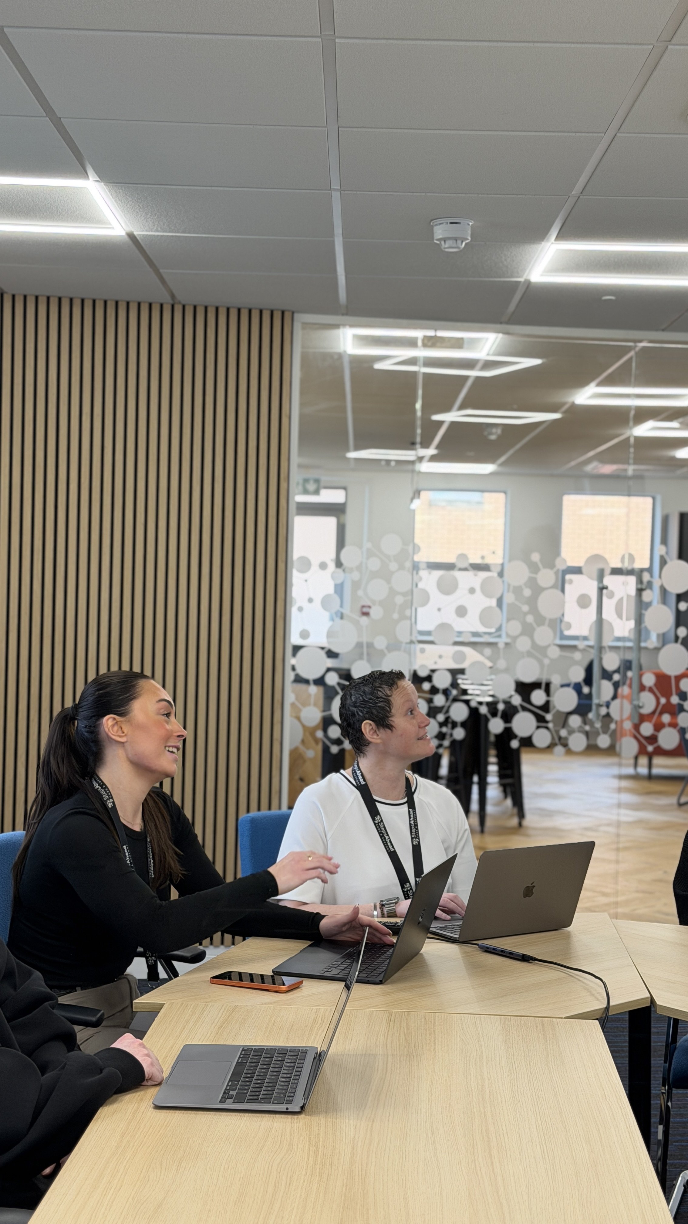 Two women sitting at a conference table with laptops, engaged in a discussion in a modern office conference room with glass wall and wooden panel decor.