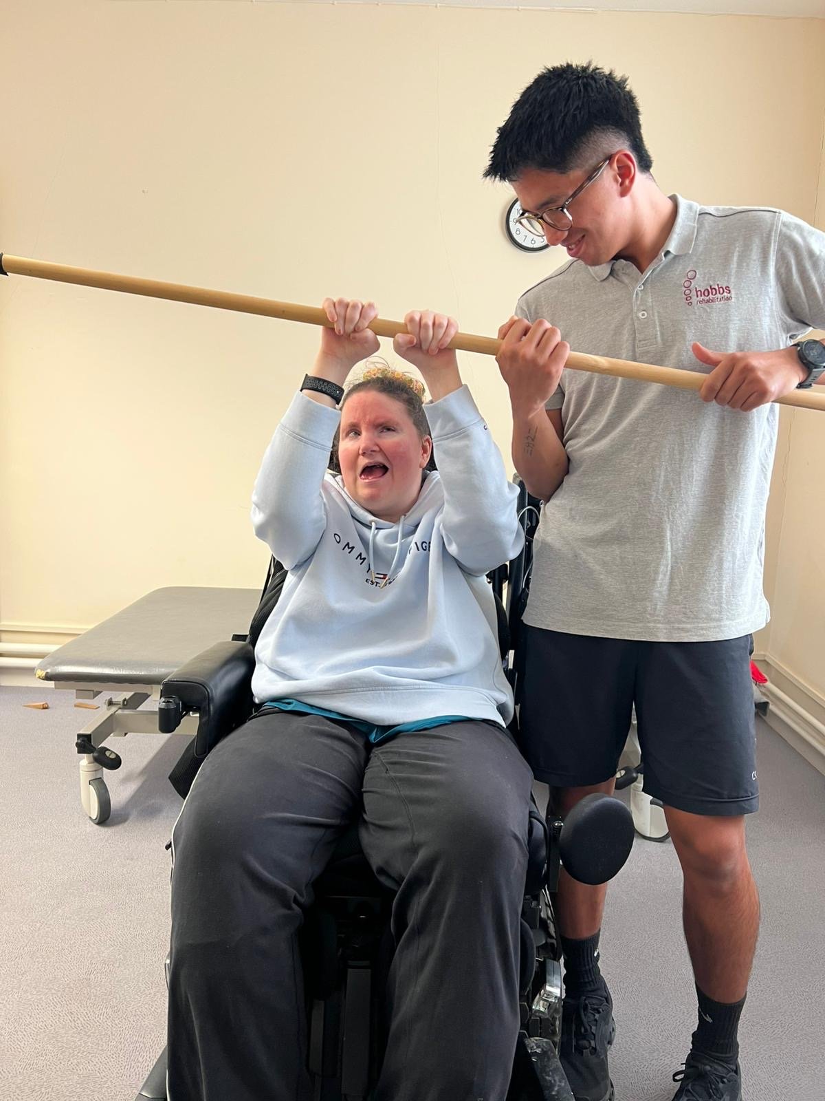 A woman in a wheelchair holding a wooden stick above her head, with a man standing next to her holding the other end of the stick, both smiling in an indoor setting.