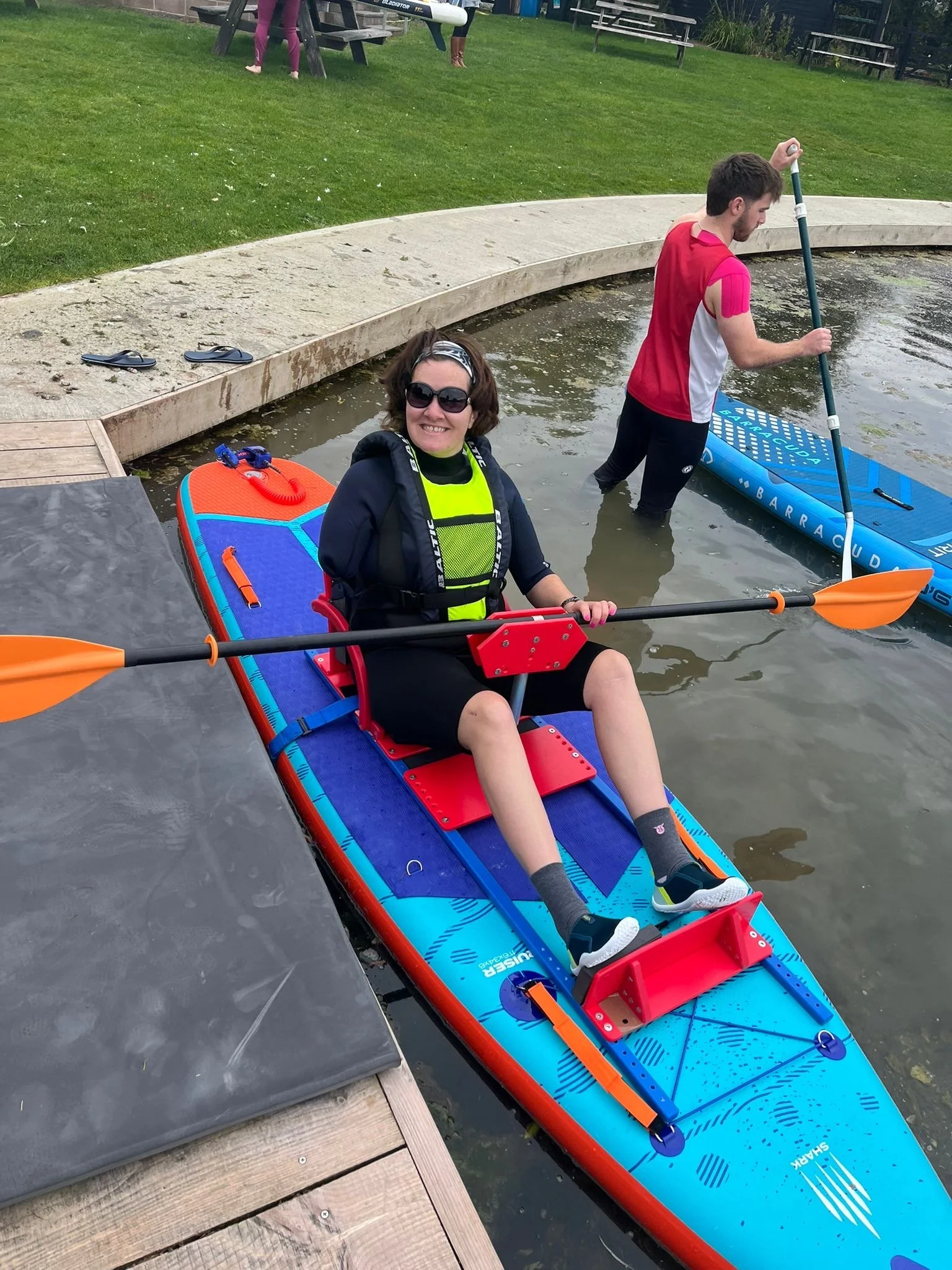 A man and woman enjoying paddleboarding at a park. The woman is sitting on an orange and blue paddleboard, smiling at the camera, wearing sunglasses, a black hoodie, and a life jacket. The man is standing in the water with a paddle, wearing a red and white shirt, and black pants. There are benches, a grassy area, and children playing in the background.