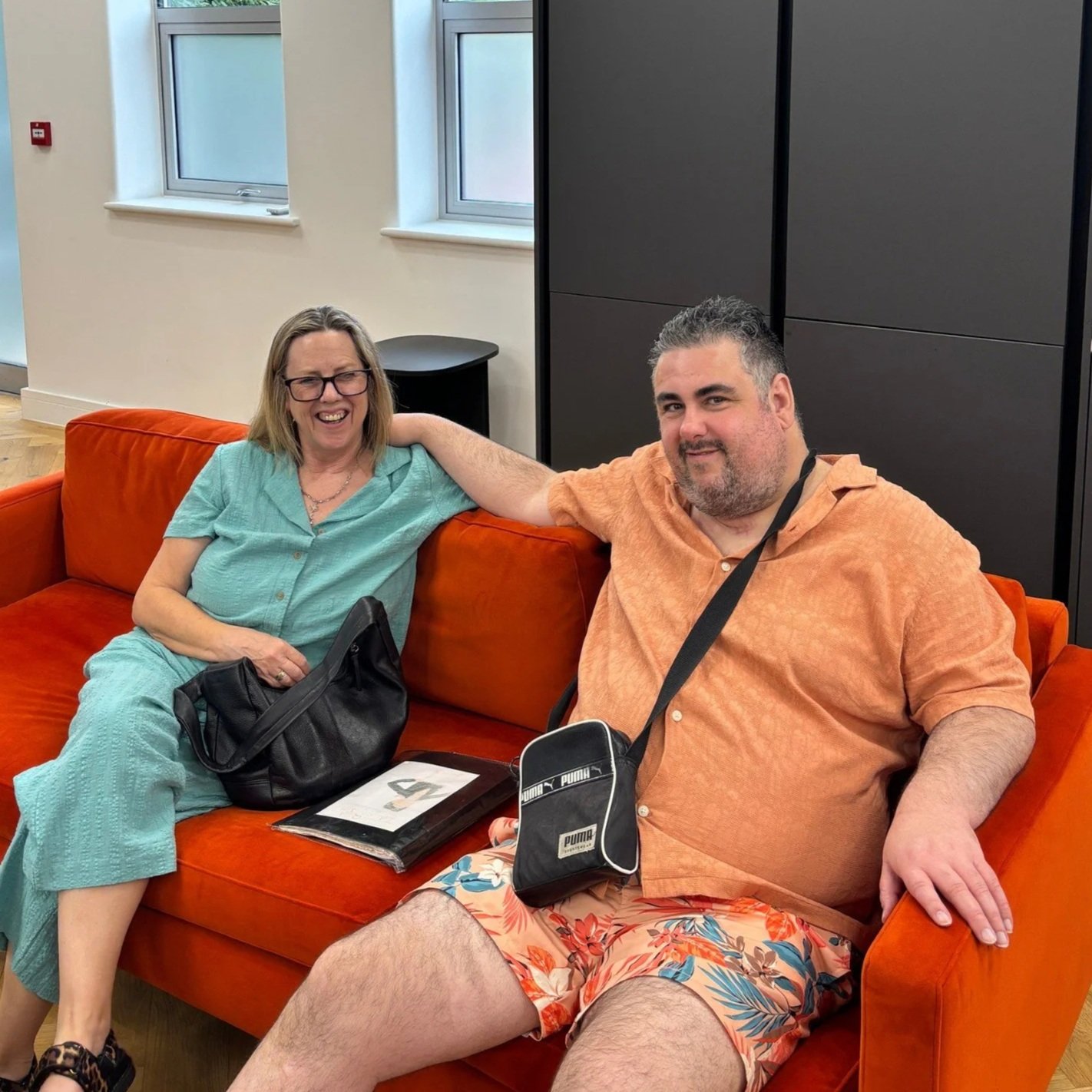 Three people sitting at a round table in a glass-walled conference room with green trees outside. They are looking at the camera, with documents and a laptop on the table.