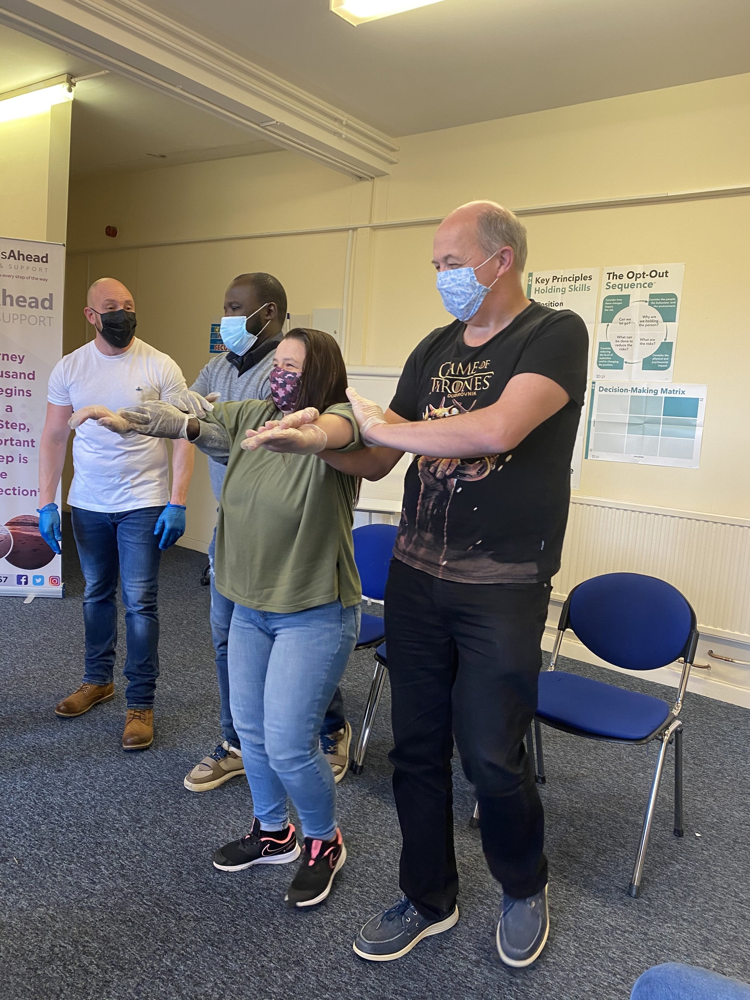 Group of five diverse people practicing hand safety measures in a classroom or training room. All are wearing face masks and gloves, standing in a line with their arms extended and palms facing down.