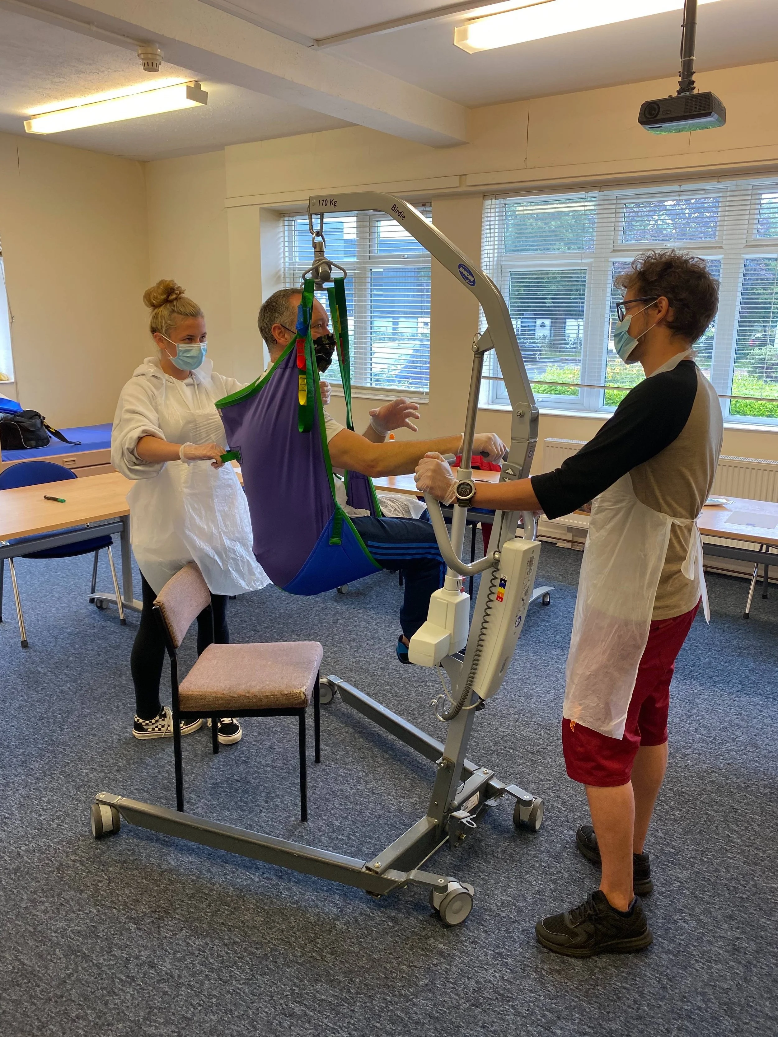 A person is demonstrating a patient lift with assistance from two healthcare workers in a room with large windows and blinds, all wearing face masks and protective gear.