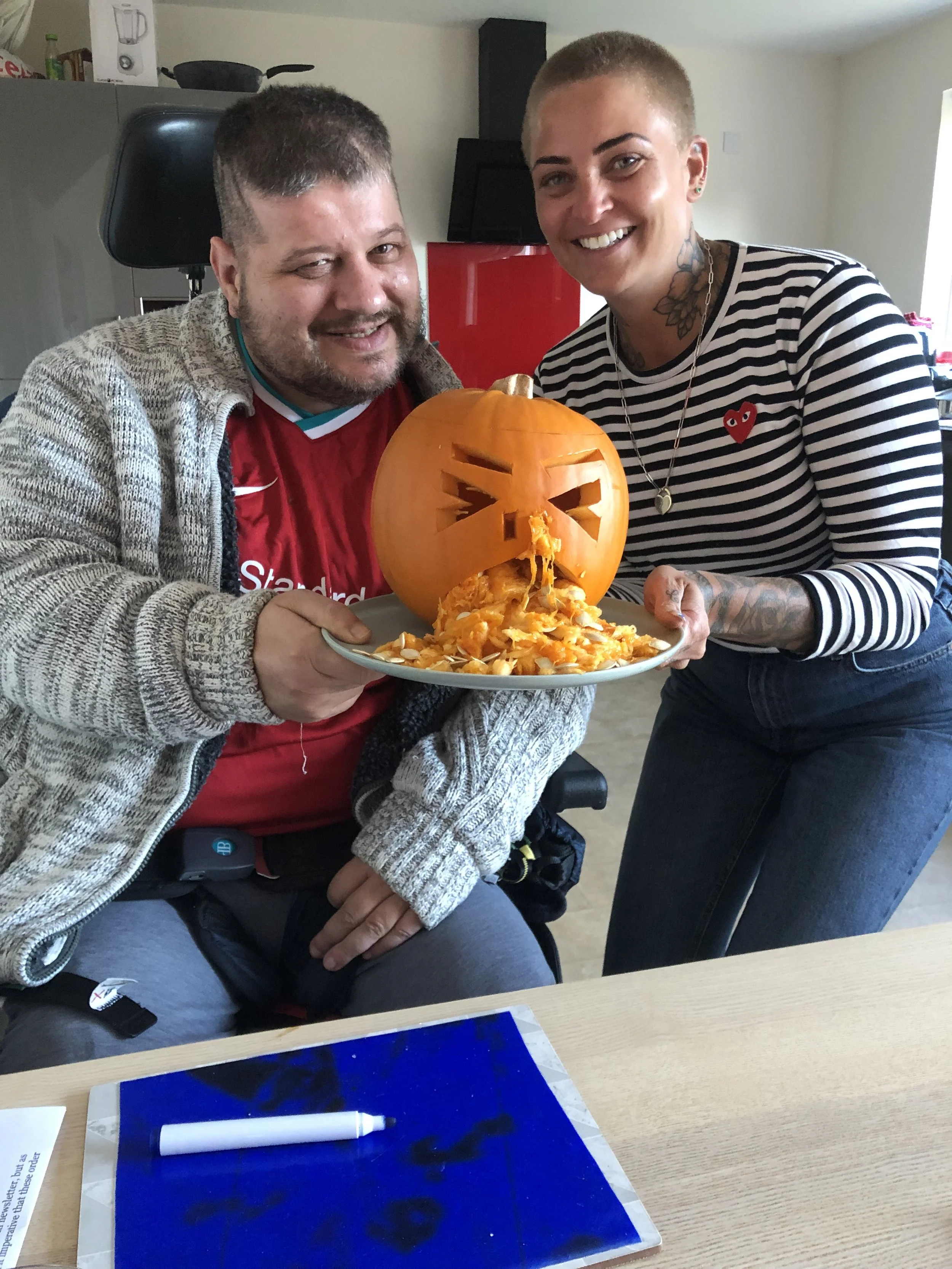A woman with dark hair in a bun uses a knife to cut a blindfolded man's head while he is seated in a motorized wheelchair indoors in front of large windows showing a backyard.