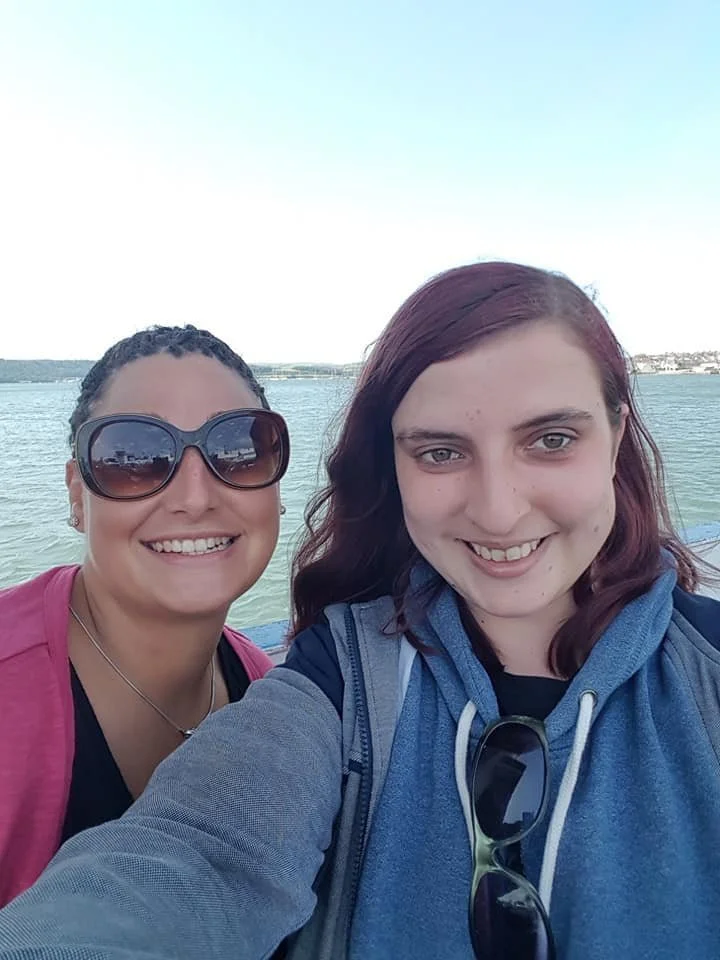 Two women taking a selfie near a body of water, with a clear sky and distant shoreline in the background.