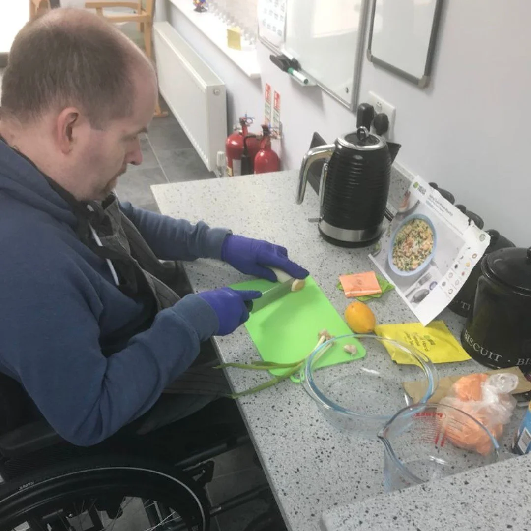 Person in a wheelchair chopping garlic on a green cutting board at a kitchen counter. The counter has a yellow lemon, a glass bowl, and a plastic bag of garlic. In the background, there are fire extinguishers, a kettle, and cooking instructions.