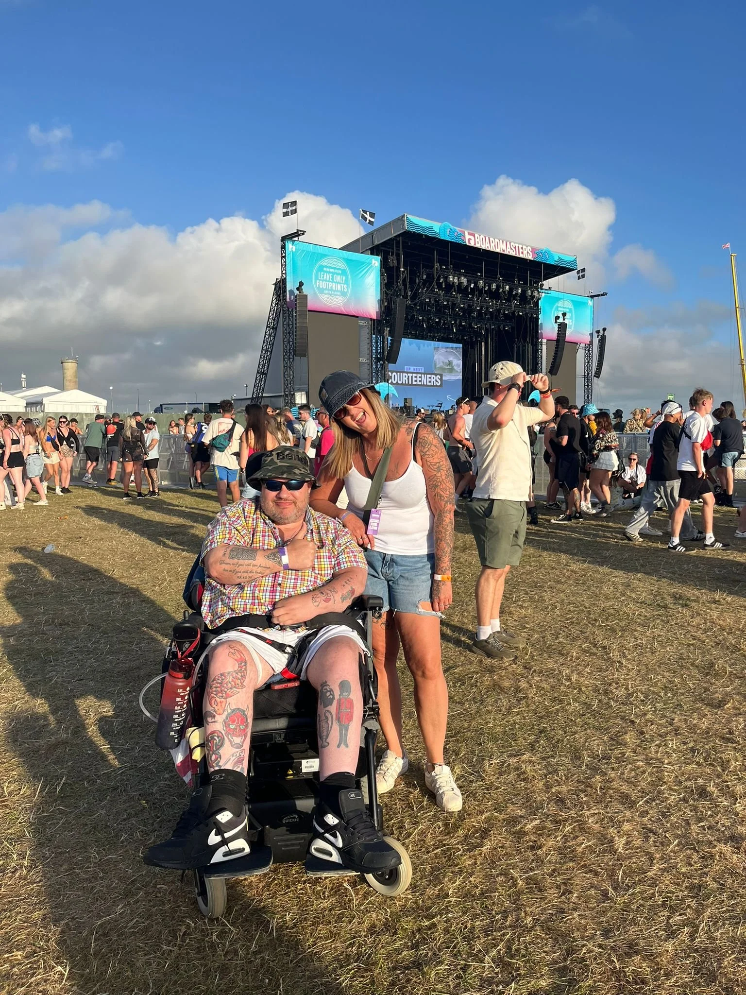 People enjoying a music festival outdoors on a sunny day, with a large stage and crowd in the background. Two women are in the foreground, one sitting in a motorized wheelchair and the other standing beside her, both smiling and posing for the photo.