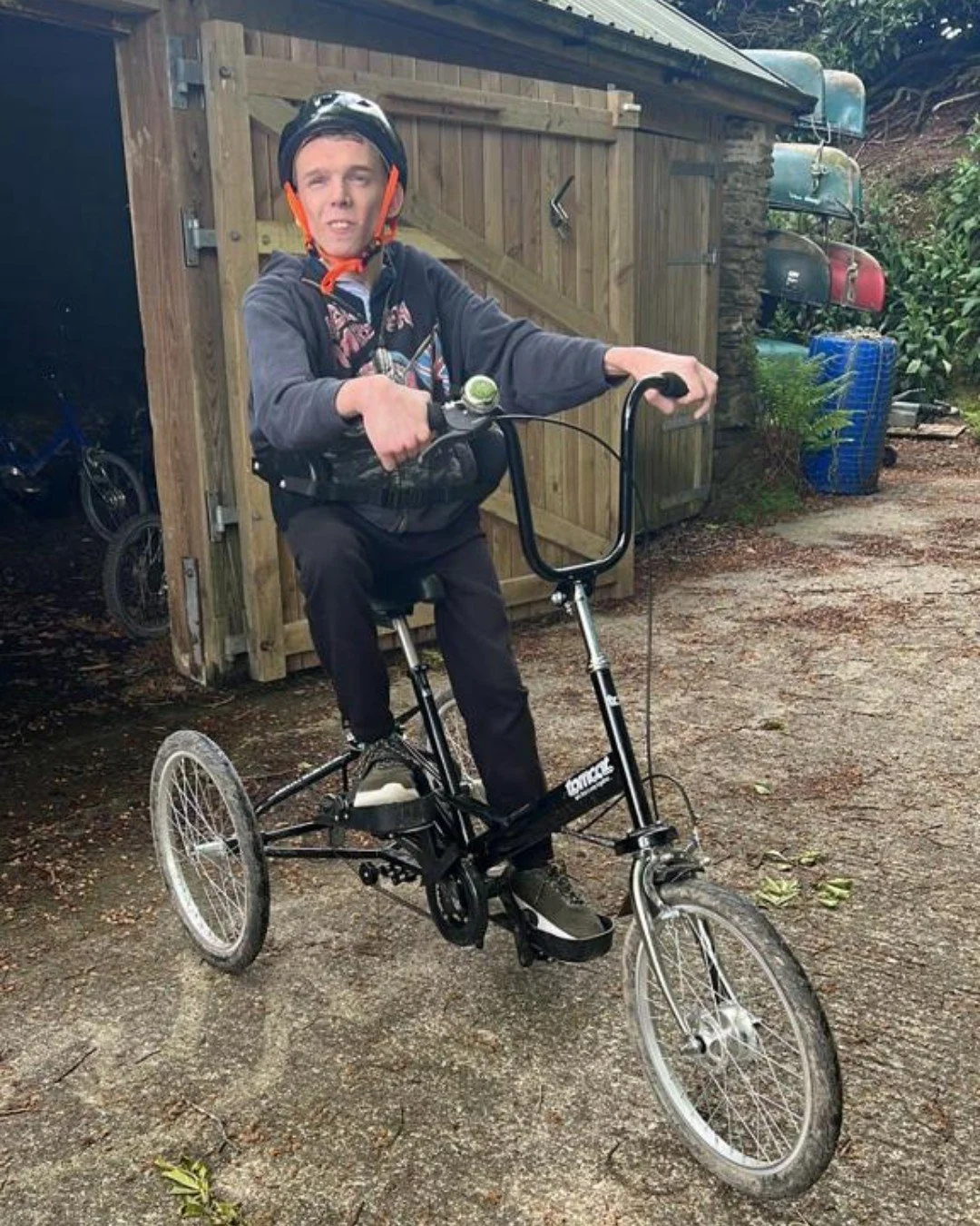 A young man wearing a helmet riding a small three-wheeled bicycle outdoors in a yard with a wooden shed and colorful canoes in the background.