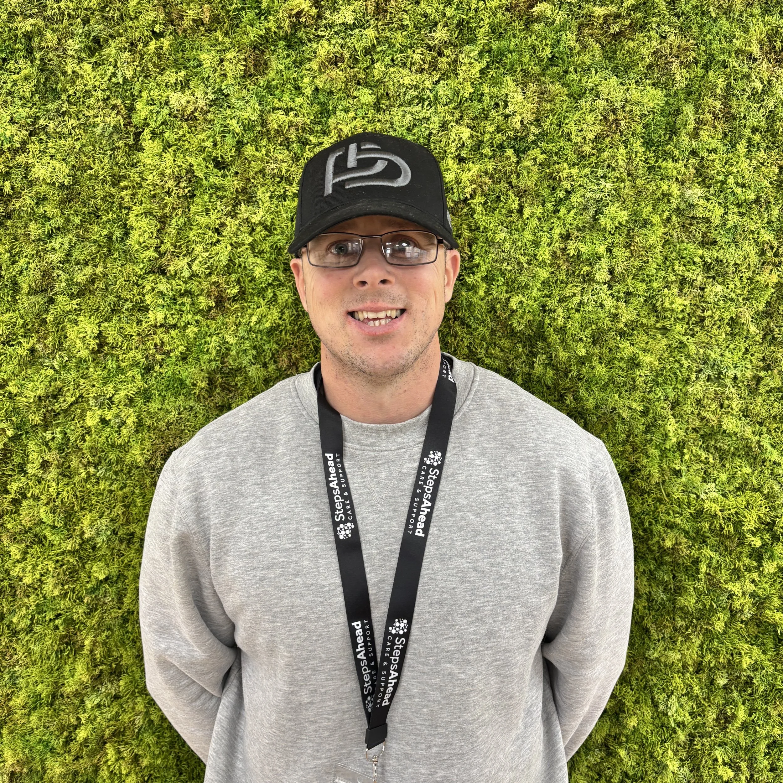 A man wearing glasses, a black cap, and a grey sweatshirt standing in front of a bright green moss wall.
