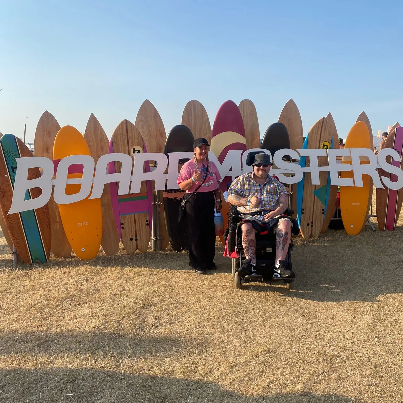 Two people posing in front of a large surfboard display with a sign that says 'Board Masters'. One person is standing and the other is sitting in a power wheelchair. The standing person has long braided hair, is smiling, and holding a water bottle. The person in the wheelchair has tattoos, a hat, and is wearing sunglasses.