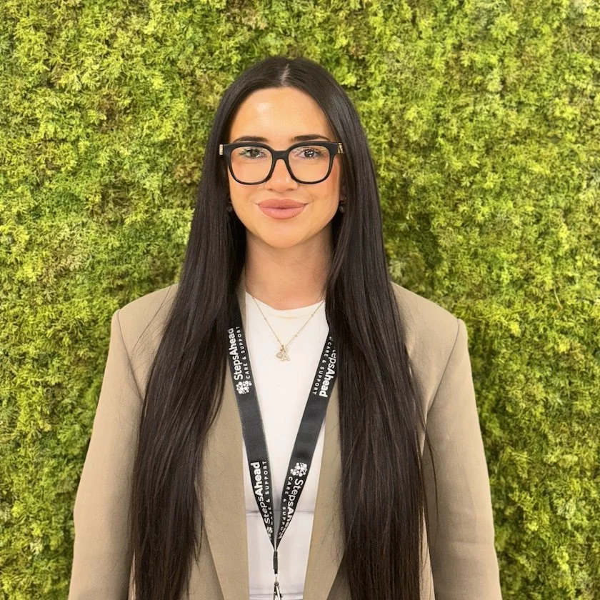 A woman with long dark hair, wearing glasses, a black lanyard, a beige blazer, and a white shirt, standing in front of a green leafy wall.