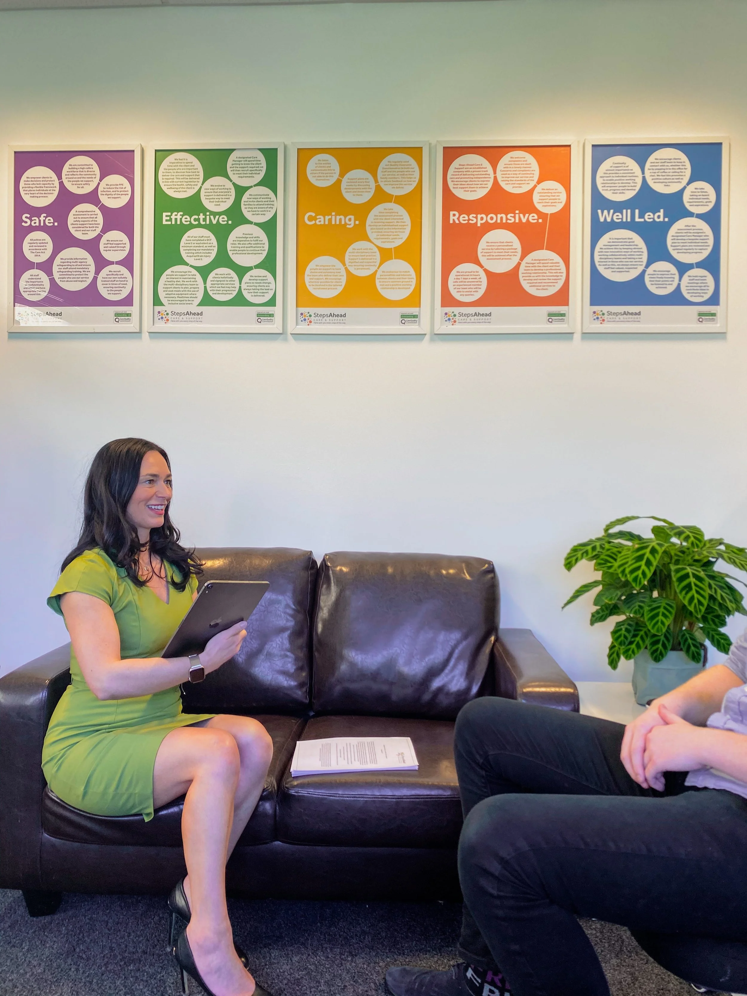 A woman in a green dress sitting on a brown leather couch, holding a tablet, and smiling during a conversation with another person whose hands are visible. Behind them, five colorful posters with words like Safe, Effective, Caring, Responsive, and Well Led are displayed on the wall, along with a potted plant.