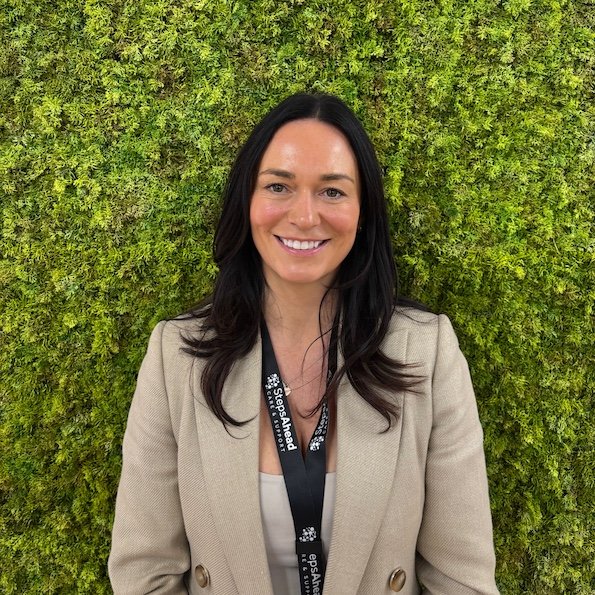 A woman with long dark hair, smiling, standing in front of a green moss wall, wearing a beige blazer and a black lanyard.