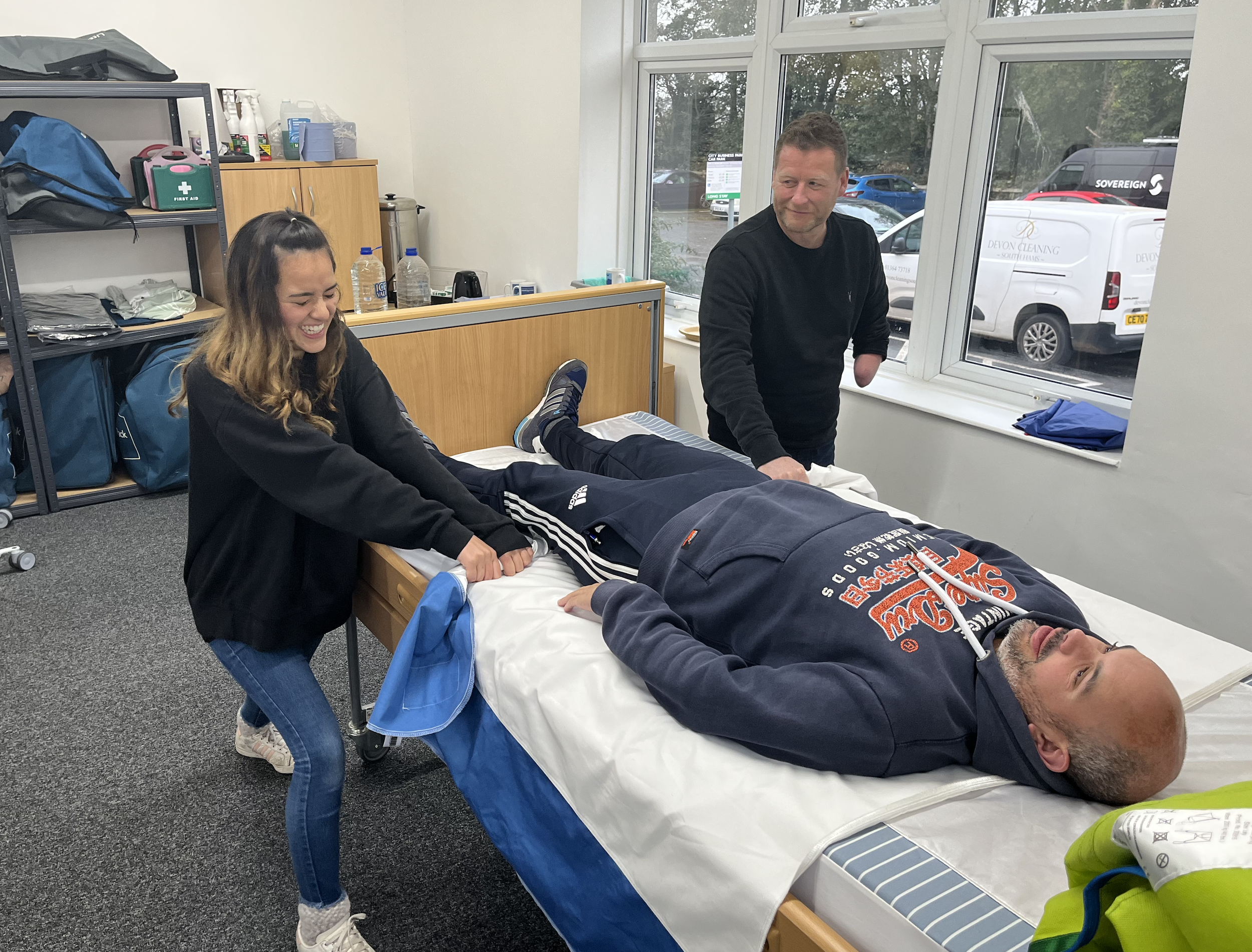 A woman and a man are on a hospital bed. The woman is smiling and pulling the man's legs, while the man is lying down and appears relaxed. A second man is standing nearby, observing them.