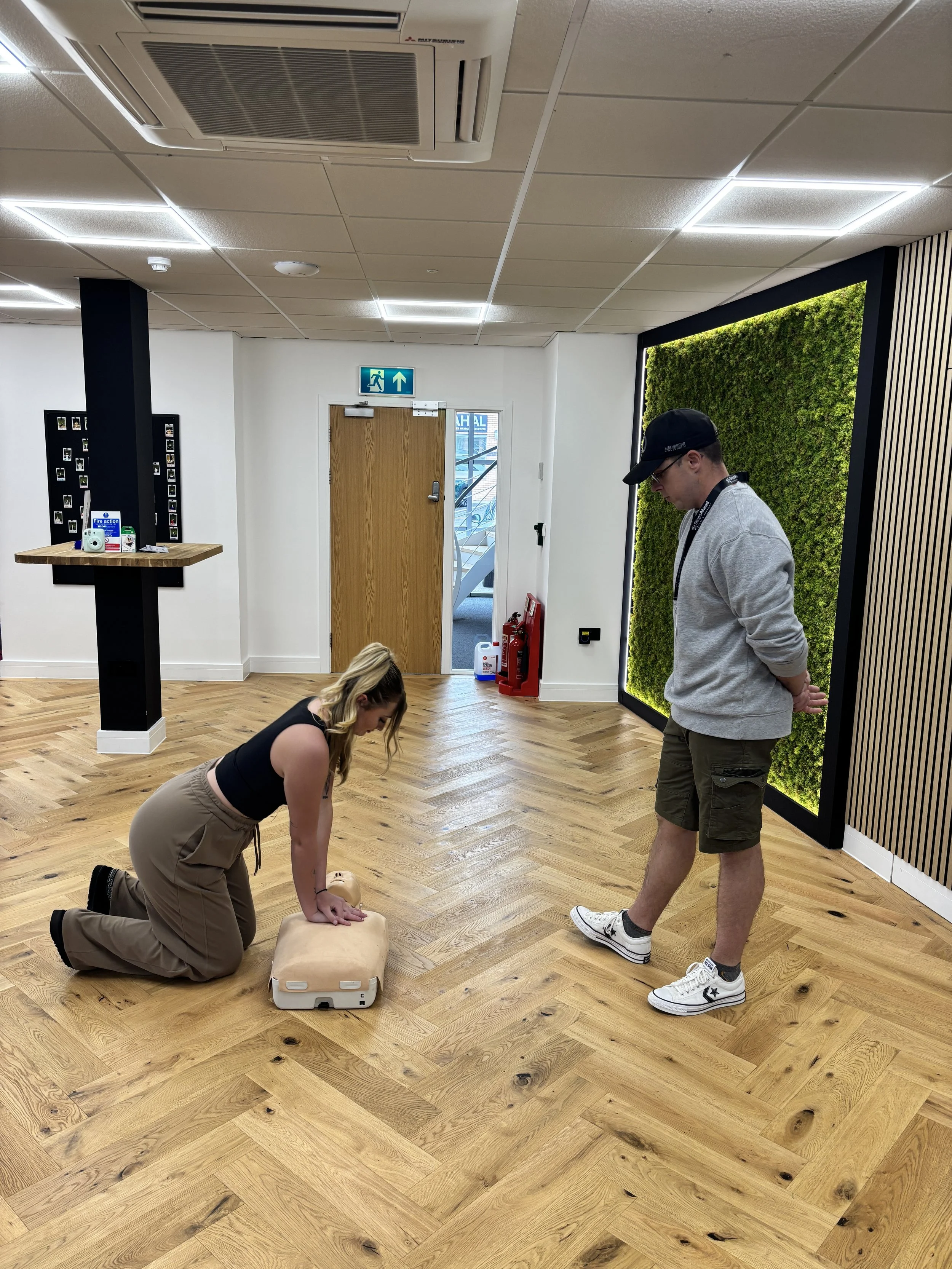 A woman practicing CPR on a mannequin while a man observes in a room with wood flooring, a green wall panel, and a fire escape door.