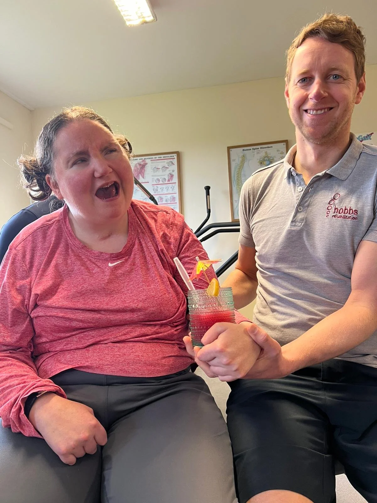 A woman in a red long-sleeve shirt and an older woman in a grey polo shirt are holding a colorful cocktail with a pink umbrella and lemon slice together. The woman in red is smiling with her mouth open, and the man in the grey shirt is smiling. They are in a room with medical posters on the wall and exercise equipment in the background.