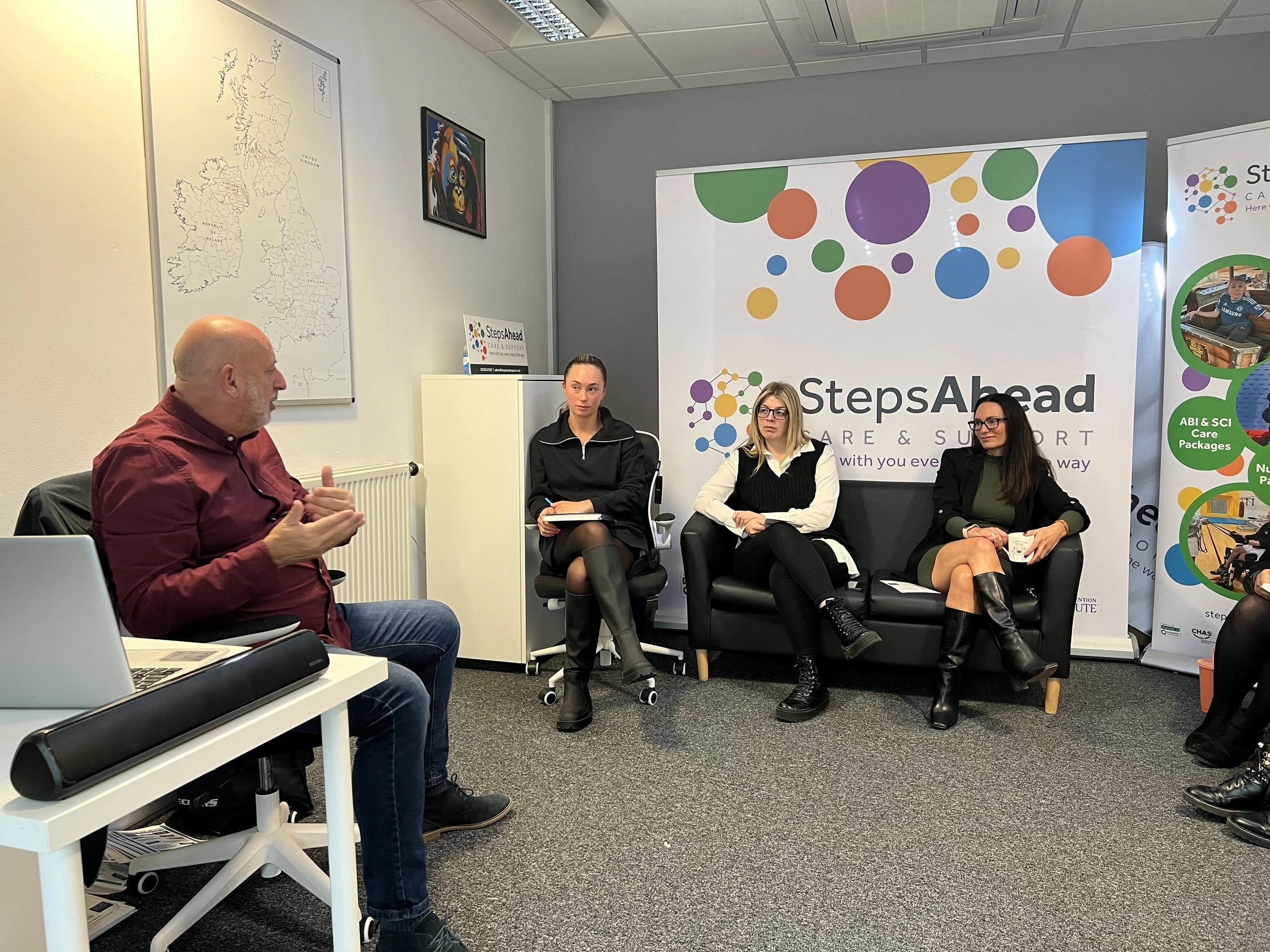 A man in a red shirt is speaking and gesturing with his hands while seated in a conference room. Across from him, three women are sitting on a black sofa, listening attentively. Behind them are two banners, one reads 'StepsAhead' with colorful logo dots, and the others have information about care packages and support programs. The room has a white wall with a map of the United Kingdom, a small colorful artwork, and the ceiling with fluorescent lights.