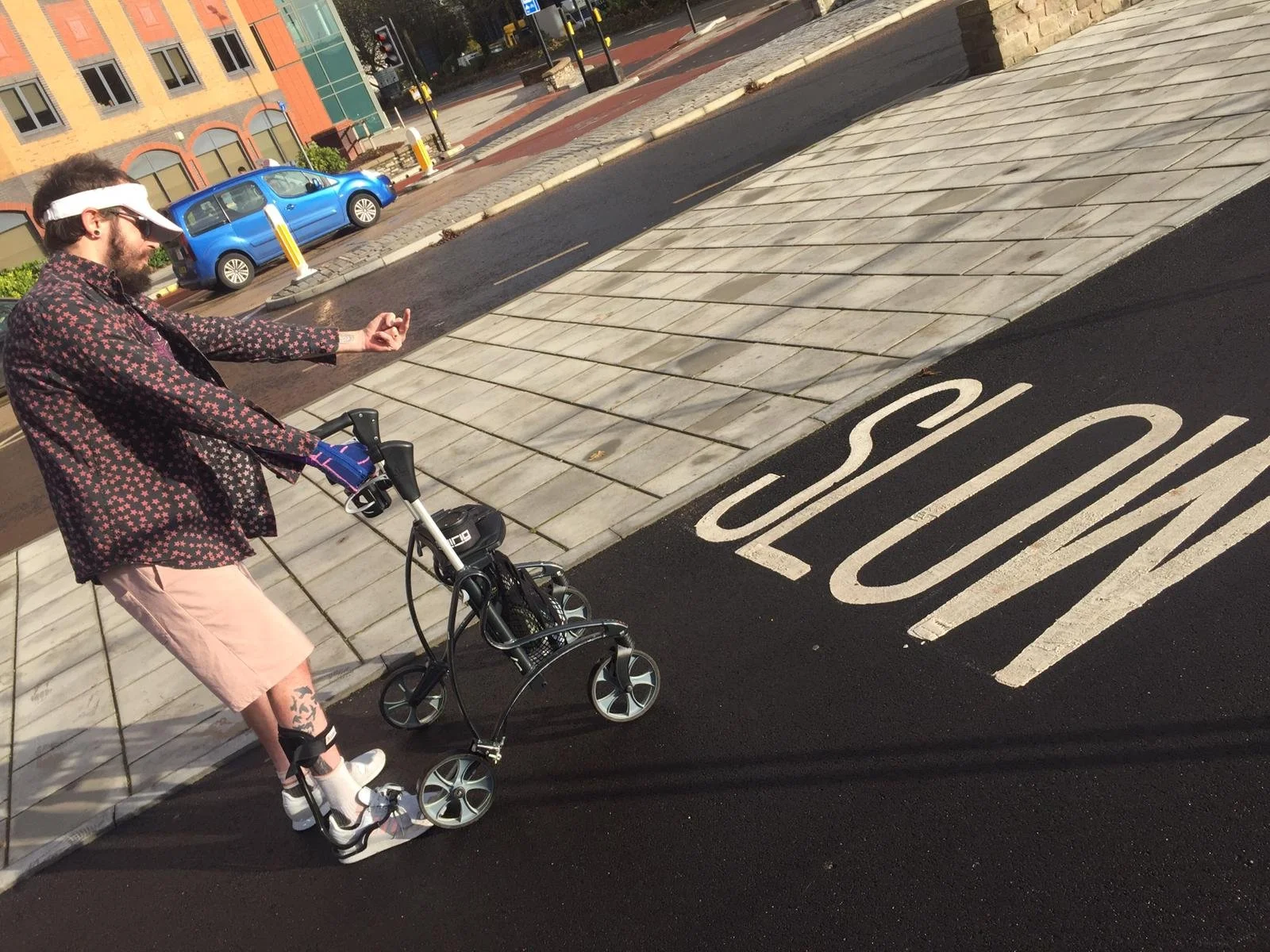 A person with a skateboard on a sidewalk near a stop sign, making an OK gesture with their hand, wearing casual clothes including a black and red patterned shirt, pink shorts, white shoes, and a white visor. There is a blue car and a building in the background.