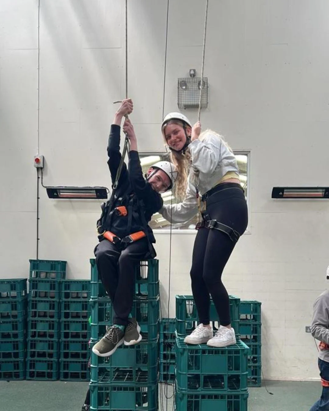Two young women with harnesses and helmets smiling and posing on stacked green crates, appearing to be participating in a team-building activity or workshop in an indoor setting.