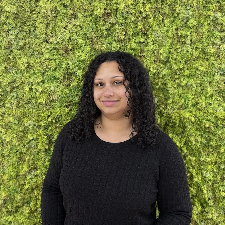 A woman with dark, curly hair, wearing a black sweater, standing in front of a lush green moss wall, smiling.