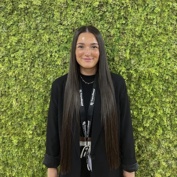 Young woman with long dark hair standing in front of a green moss wall, smiling, wearing a black blazer and a lanyard.