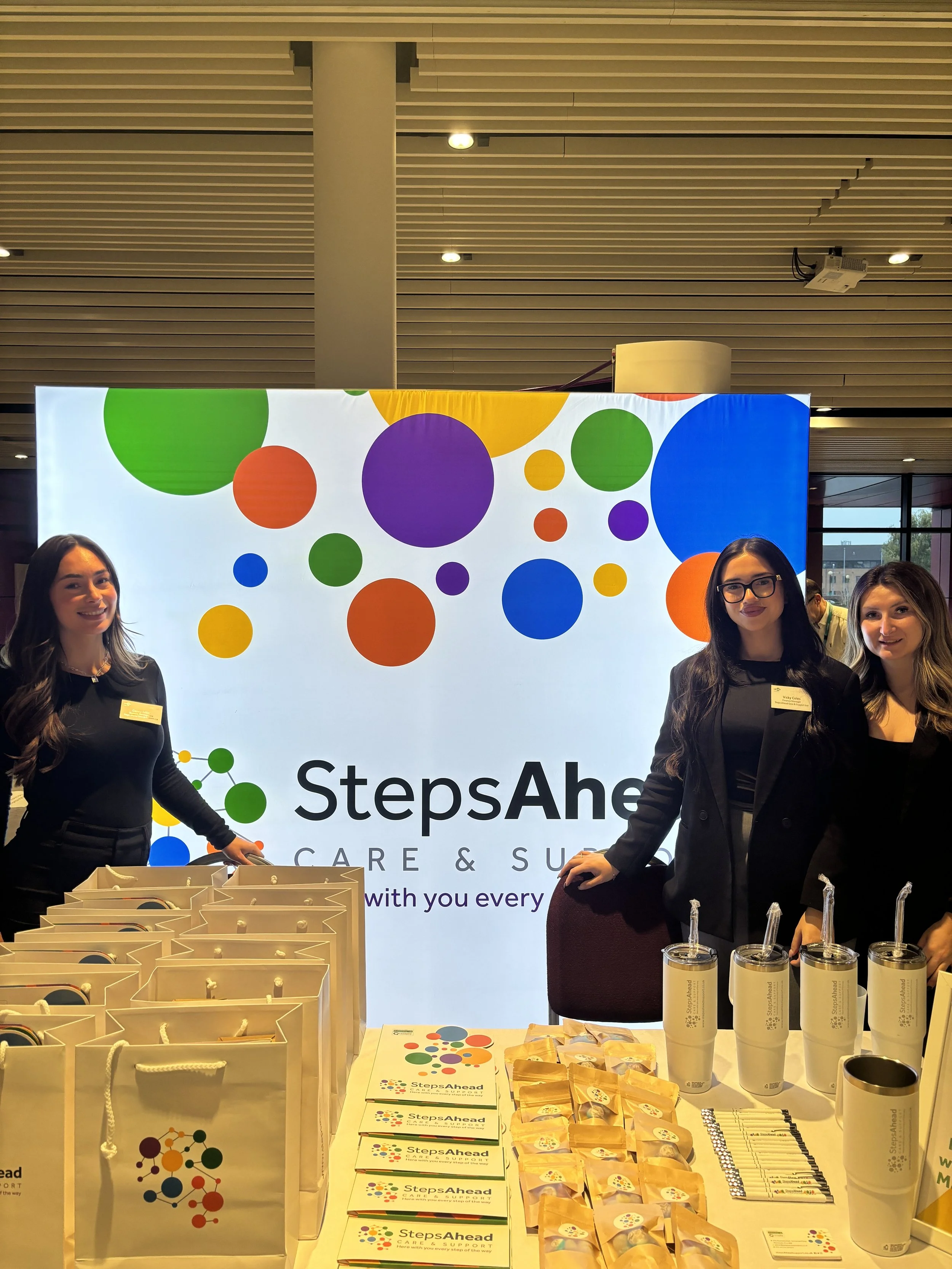 Three women are standing behind a table at a conference booth with a large illuminated sign in the background that reads "Steps Ahead Care & Support" with a colorful circle design. The table has branded bags, pamphlets, snacks, and promotional items.