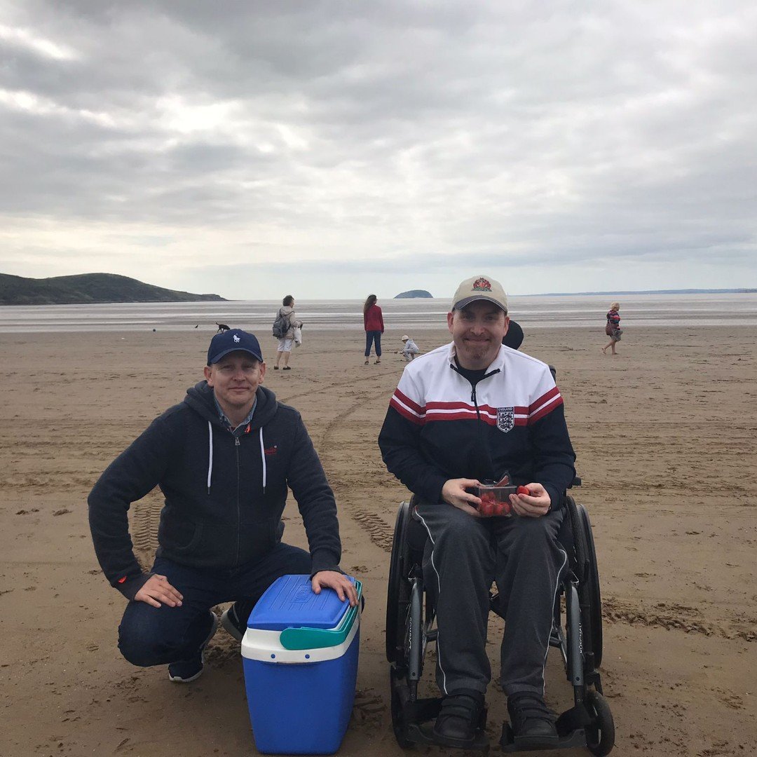 Two men on a beach, one in a wheelchair holding strawberries, the other kneeling next to a cooler. Several people in the background near the shoreline, cloudy sky above.
