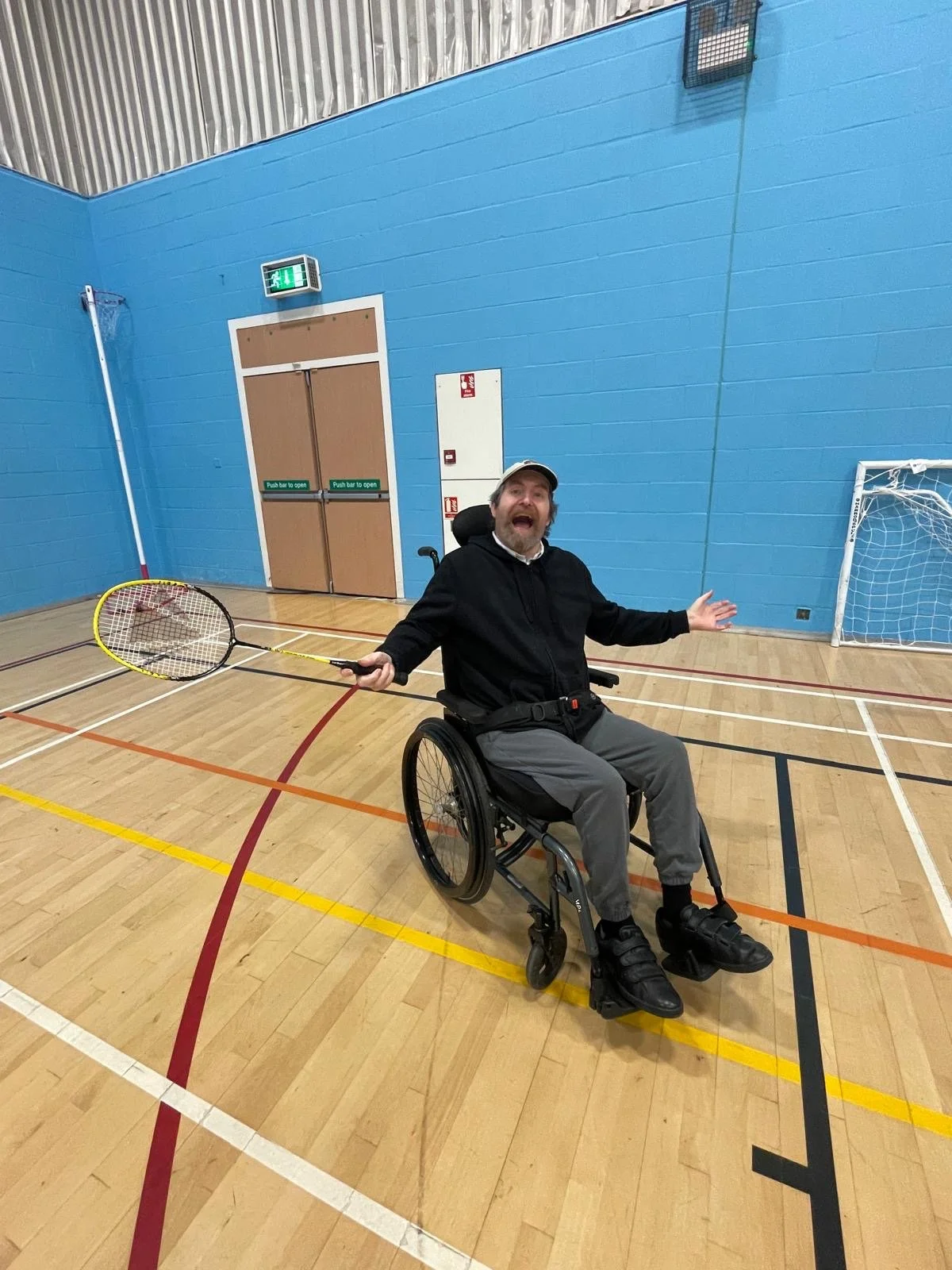 A man in a wheelchair holding a badminton racket inside an indoor sports hall with blue walls and a wooden floor.