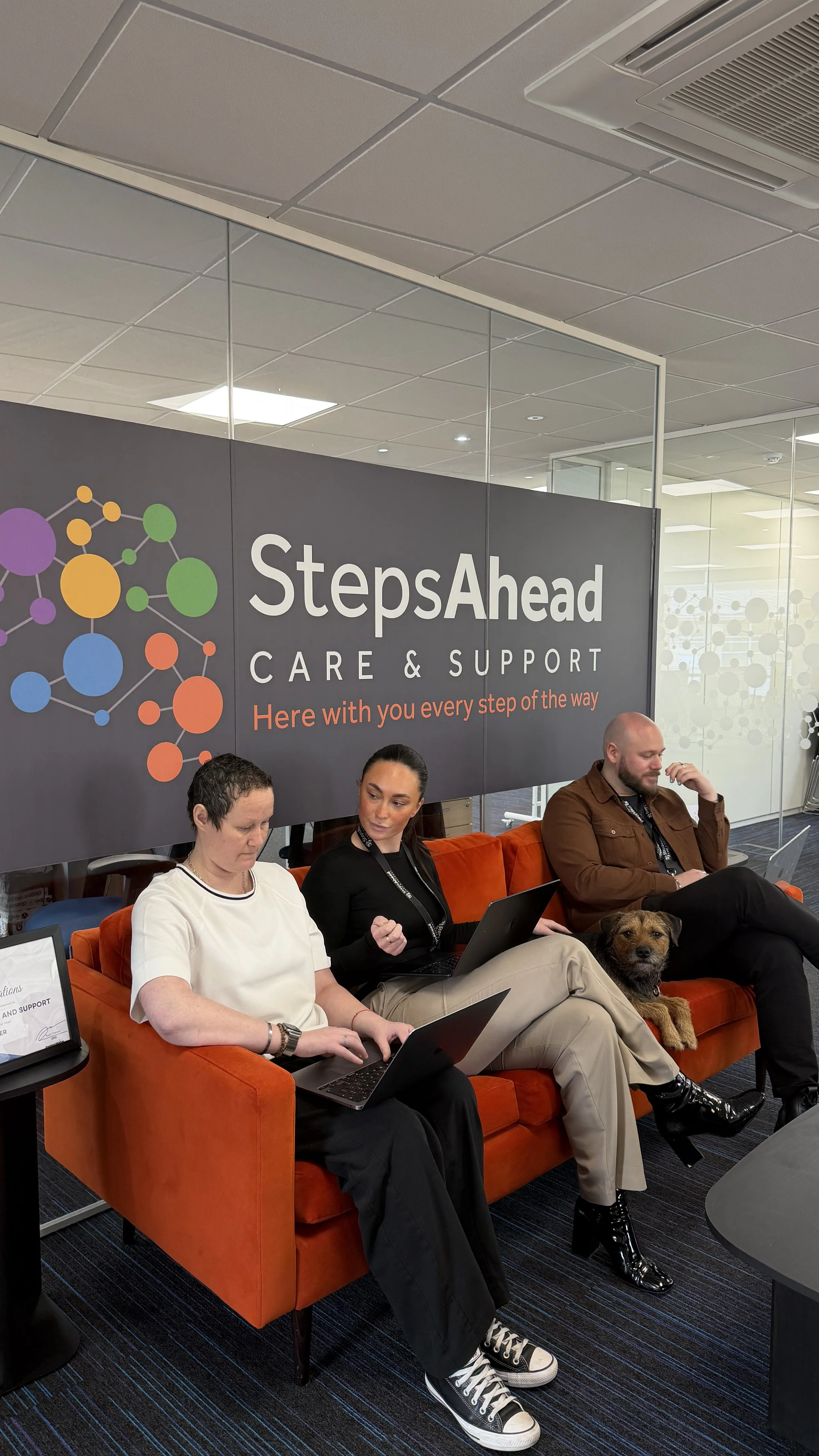 A woman with black hair, wearing a green dress, sitting on a brown leather couch, holding a tablet and smiling.