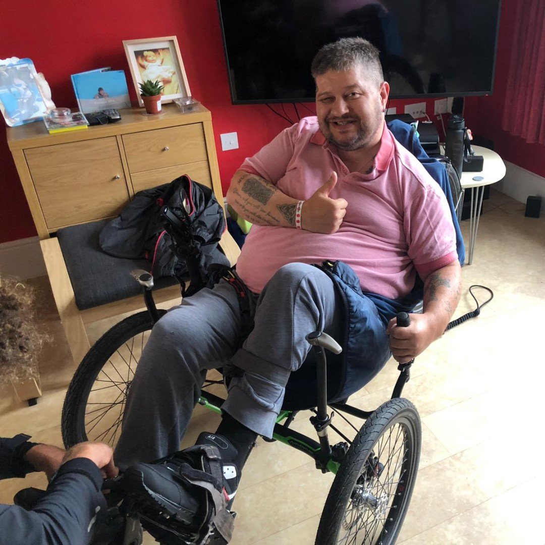 A man sitting in a wheelchair inside a room. He is giving a thumbs-up and smiling, wearing a pink polo shirt and gray pants. The room has a wooden dresser, a television, and personal items on the dresser.