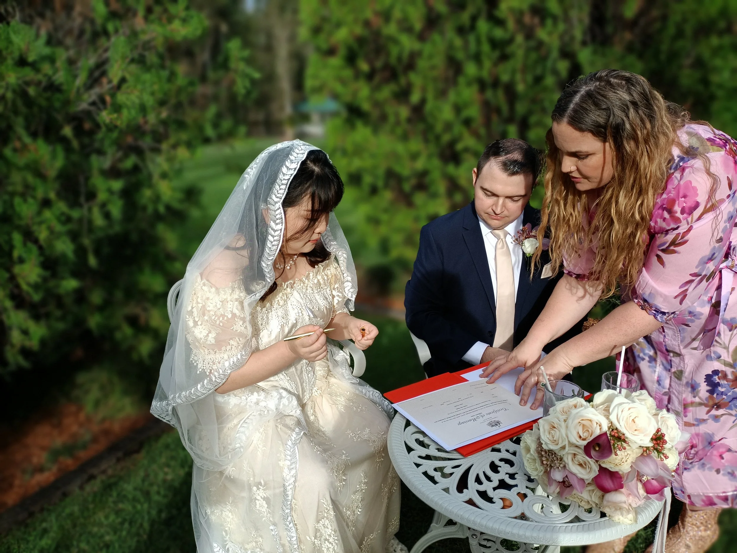 Newlyweds signing their marriage paperwork at a garden table while Maddie Hayes, Gold Coast Hinterland celebrant, guides them