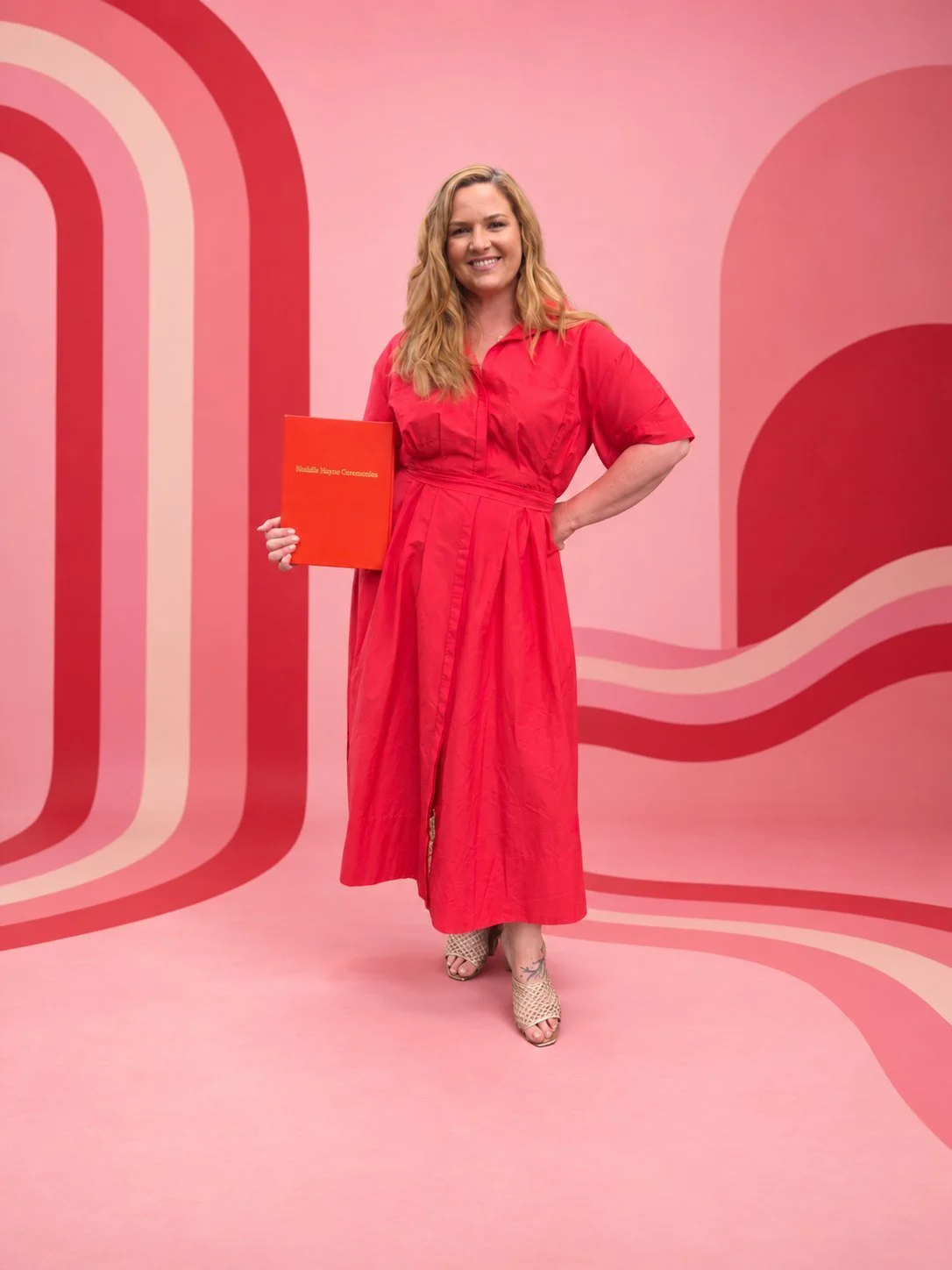 Maddie Hayes, Gold Coast Hinterland marriage celebrant, in a red dress holding her ceremony folder in front of a 70s pink and red striped backdrop