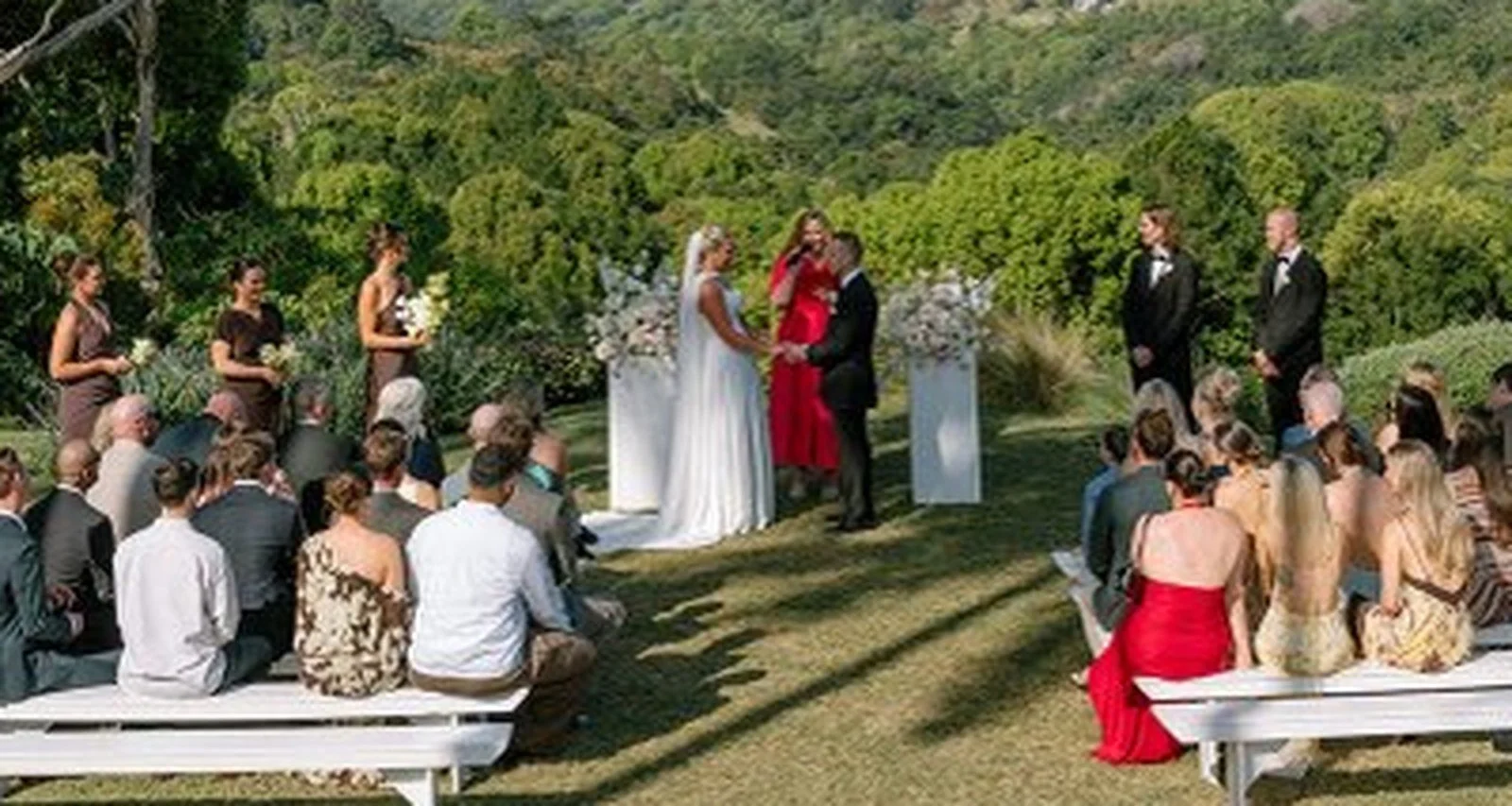 Maddie Hayes marrying a couple on a hilltop lawn in the Gold Coast Hinterland, with guests seated on white benches and green hills behind