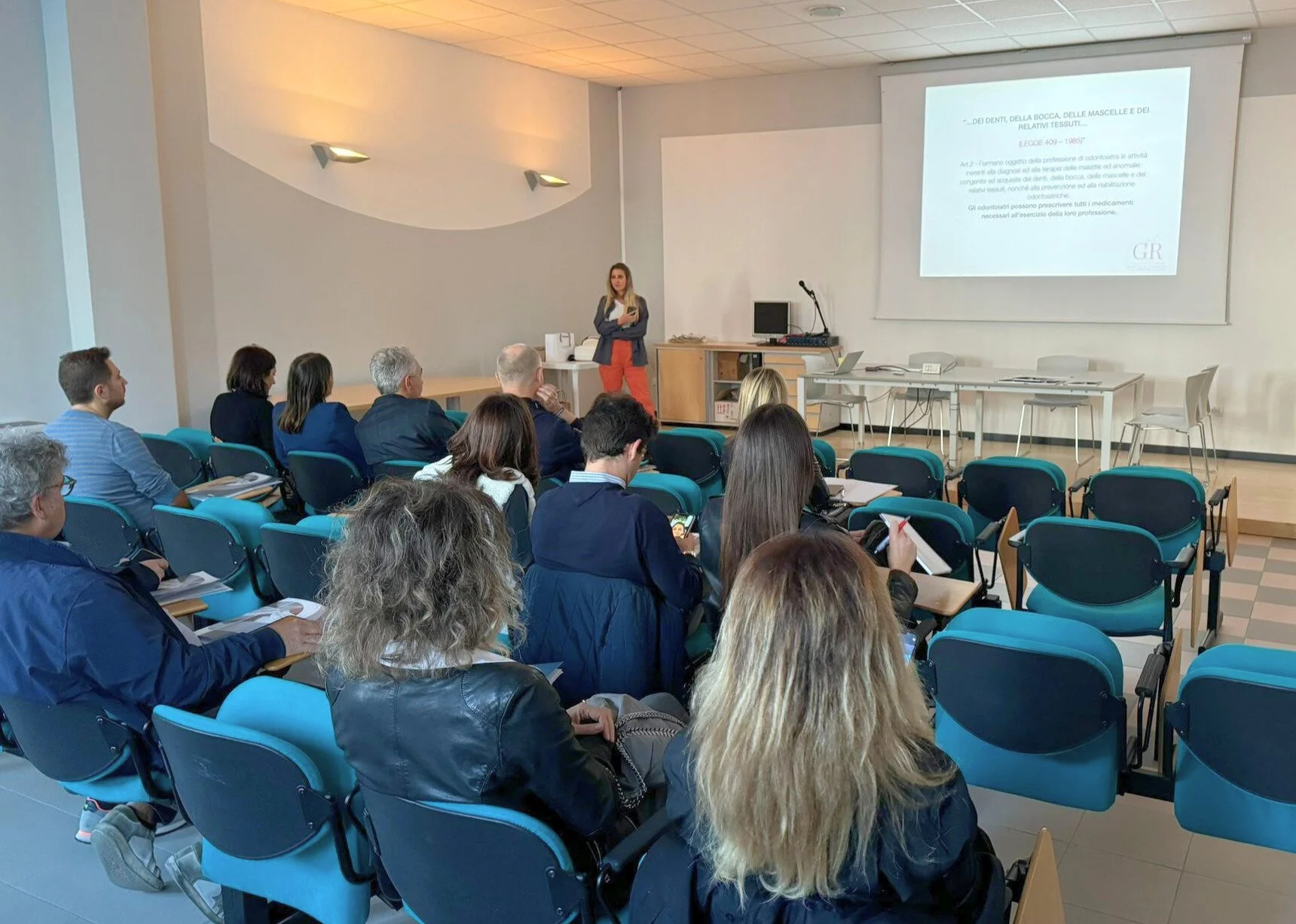 Una presentazione o conferenza si svolge in una stanza con un pubblico seduto che ascolta una donna alla presentazione, sullo schermo un testo scritto in italiano.
