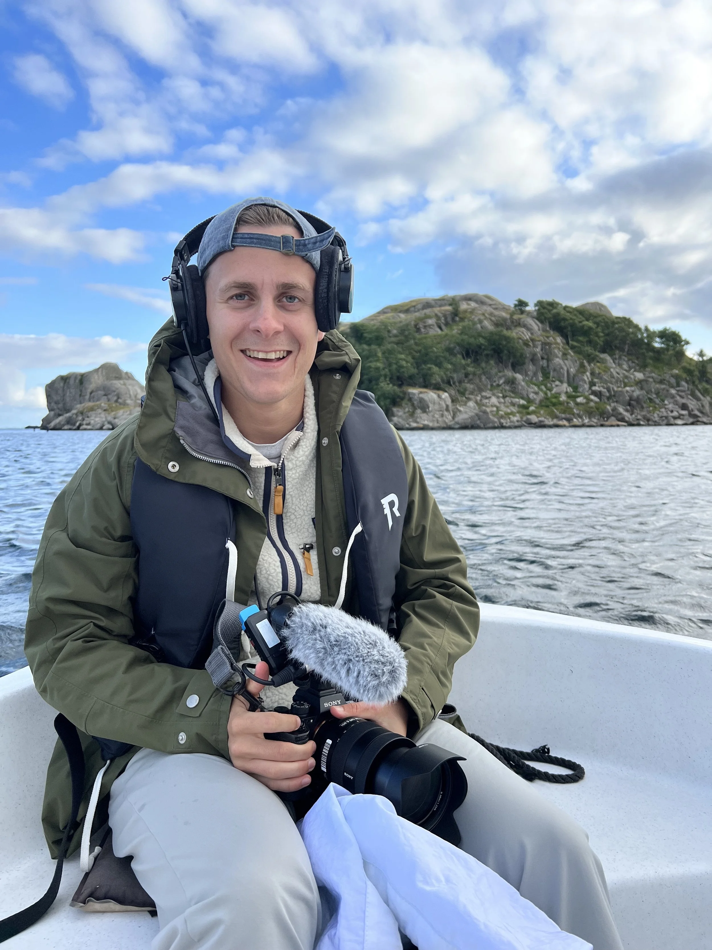 En mann som sitter i en båt med kamera og lydutstyr, omgitt av vann og øyer med steinete og grønne omgivelser.