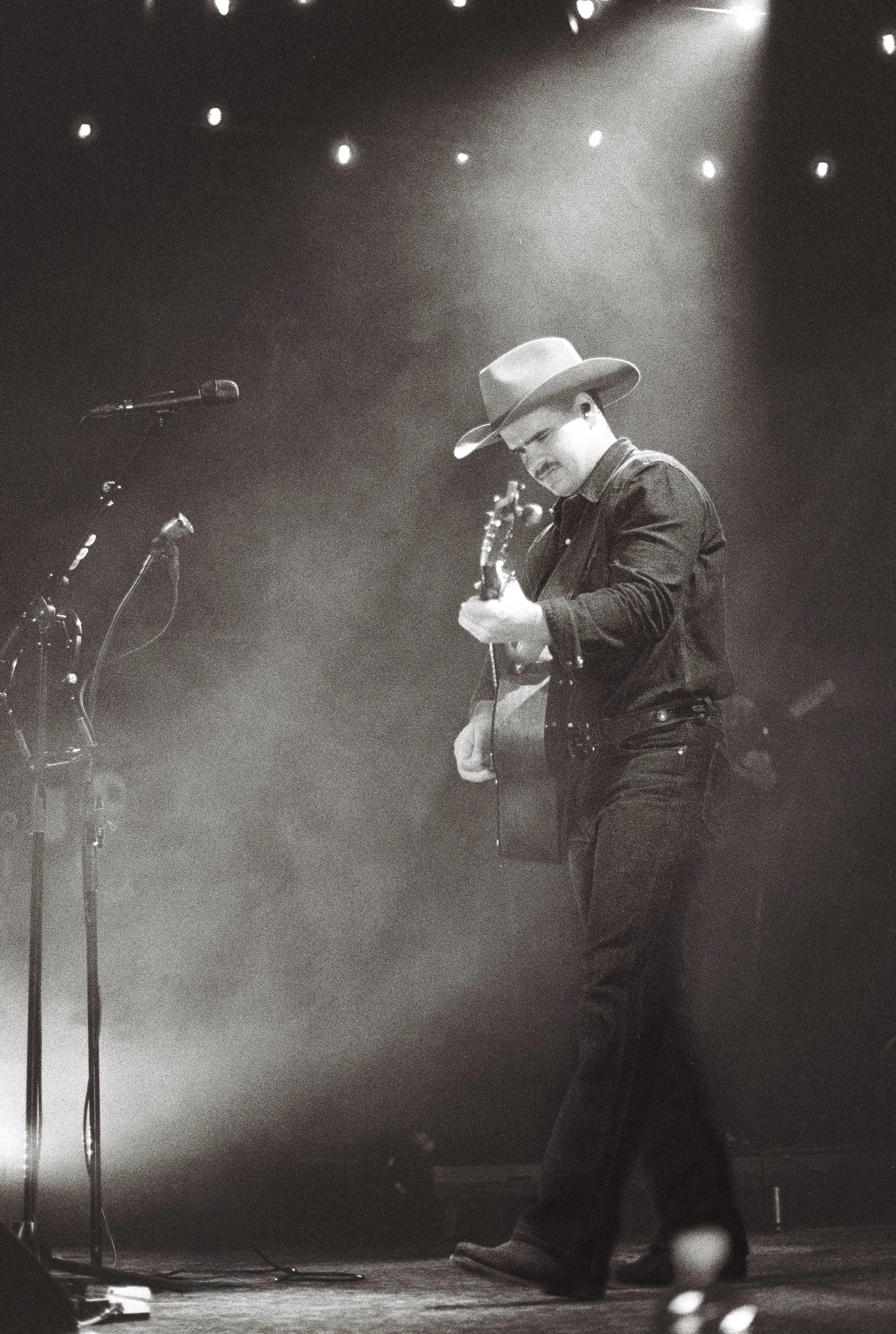 A man playing an acoustic guitar on stage, wearing a cowboy hat, glasses, and a dark jacket, with a microphone stand nearby and stage lights above.