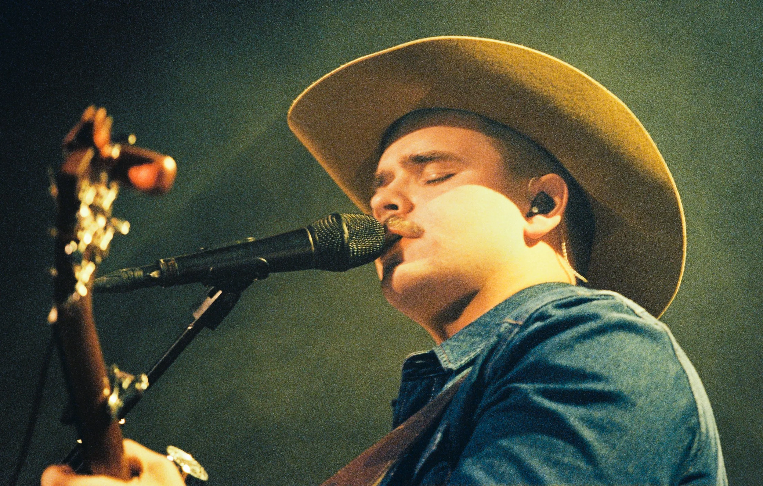 A man wearing a wide-brimmed hat and denim jacket singing into a microphone while playing a guitar.